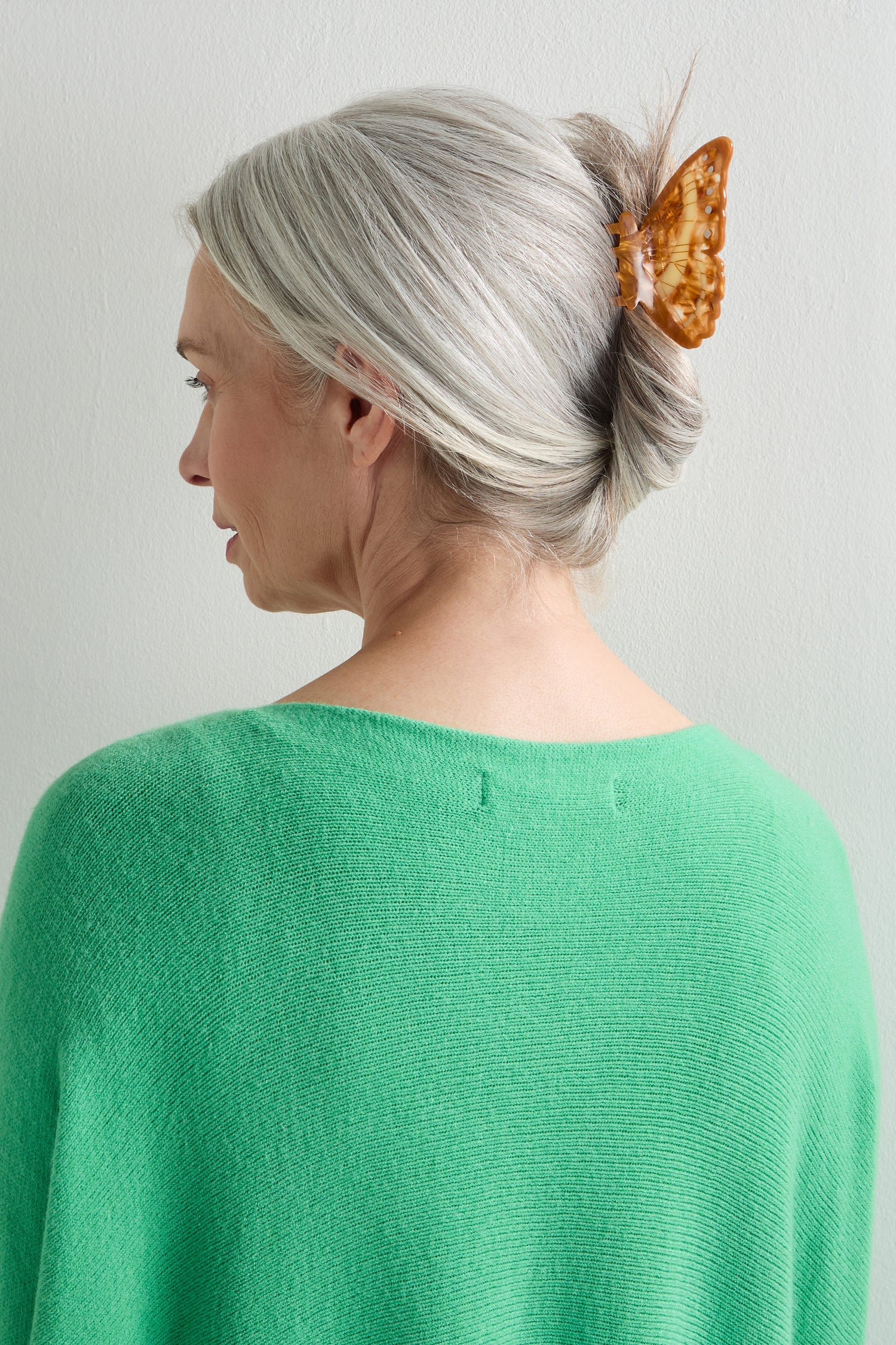 A woman with gray hair in a bun, secured by a Hand-Painted Morpho Butterfly Hair Clip, wears a green top and is seen from behind against a plain light background.