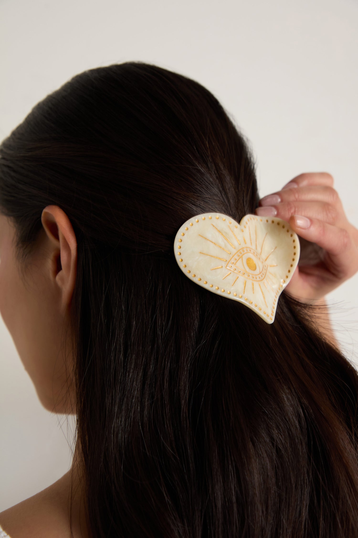 A person with long dark hair wears the Hand Painted Celestial Heart Hair Clip, a cream-colored accessory featuring an eye illustration and stitched details.