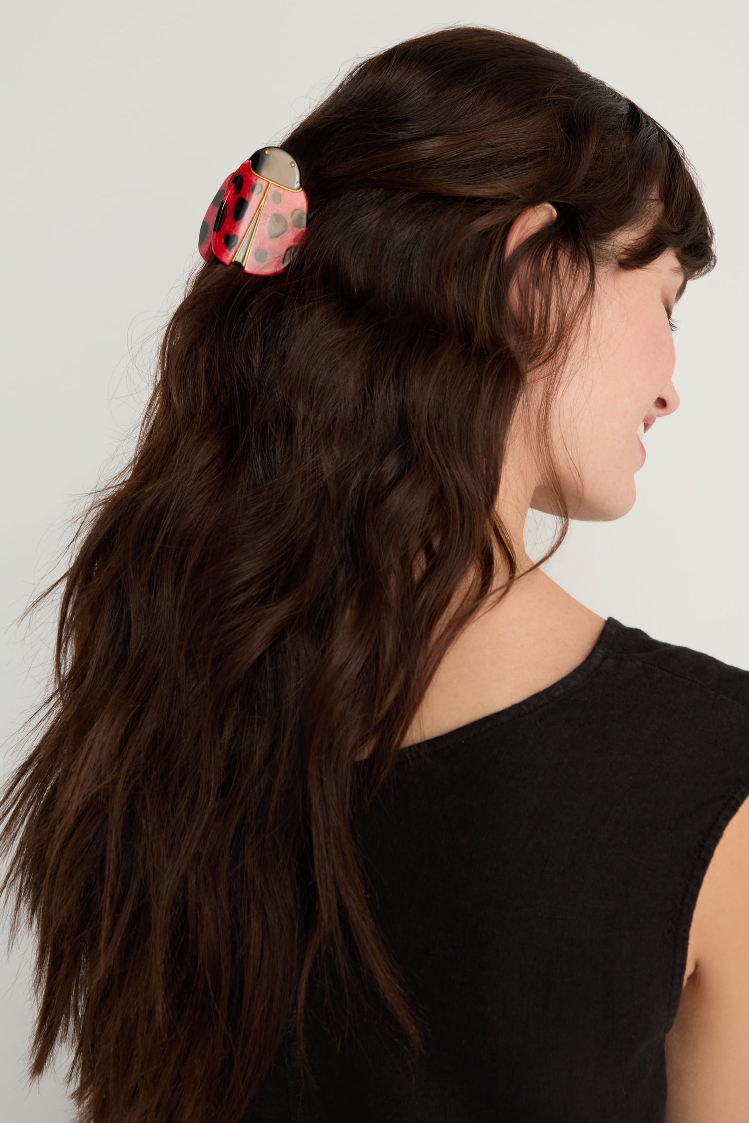 A woman with long, wavy brown hair styled half up wears a black sleeveless top and faces sideways, her hair secured with the Hand-Painted Ladybird Hair Clip.