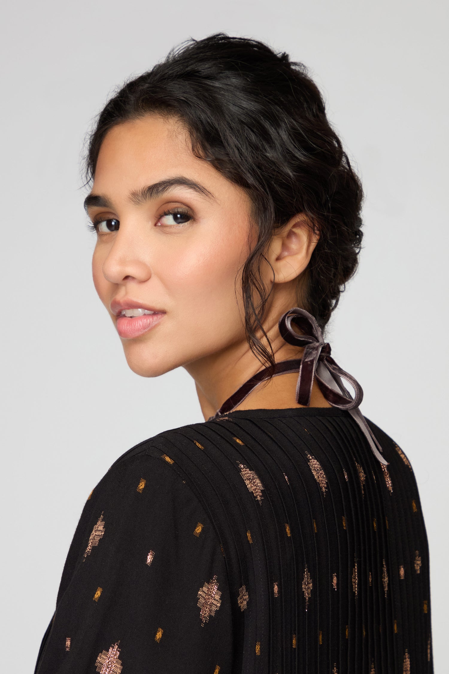 A woman with dark hair in an updo wears the Velvet Ribbon Statement Necklace tied at the back, paired with a black patterned top, as she looks over her shoulder against a plain background.