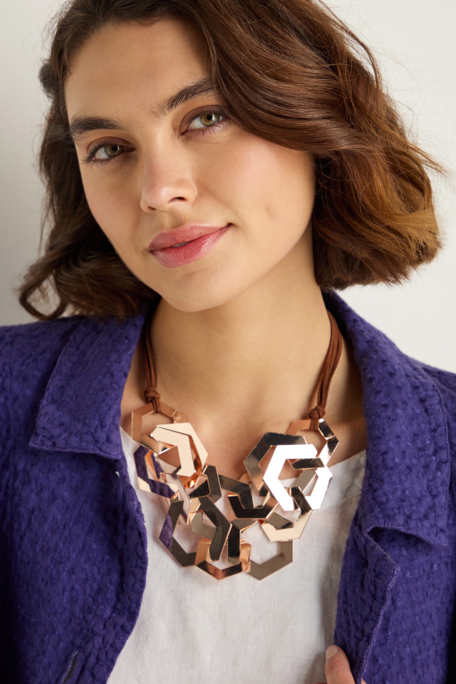 Woman with wavy brown hair looks at the camera, wearing a purple textured jacket, white top, and the Hexagon Links Necklace in metallic tones.
