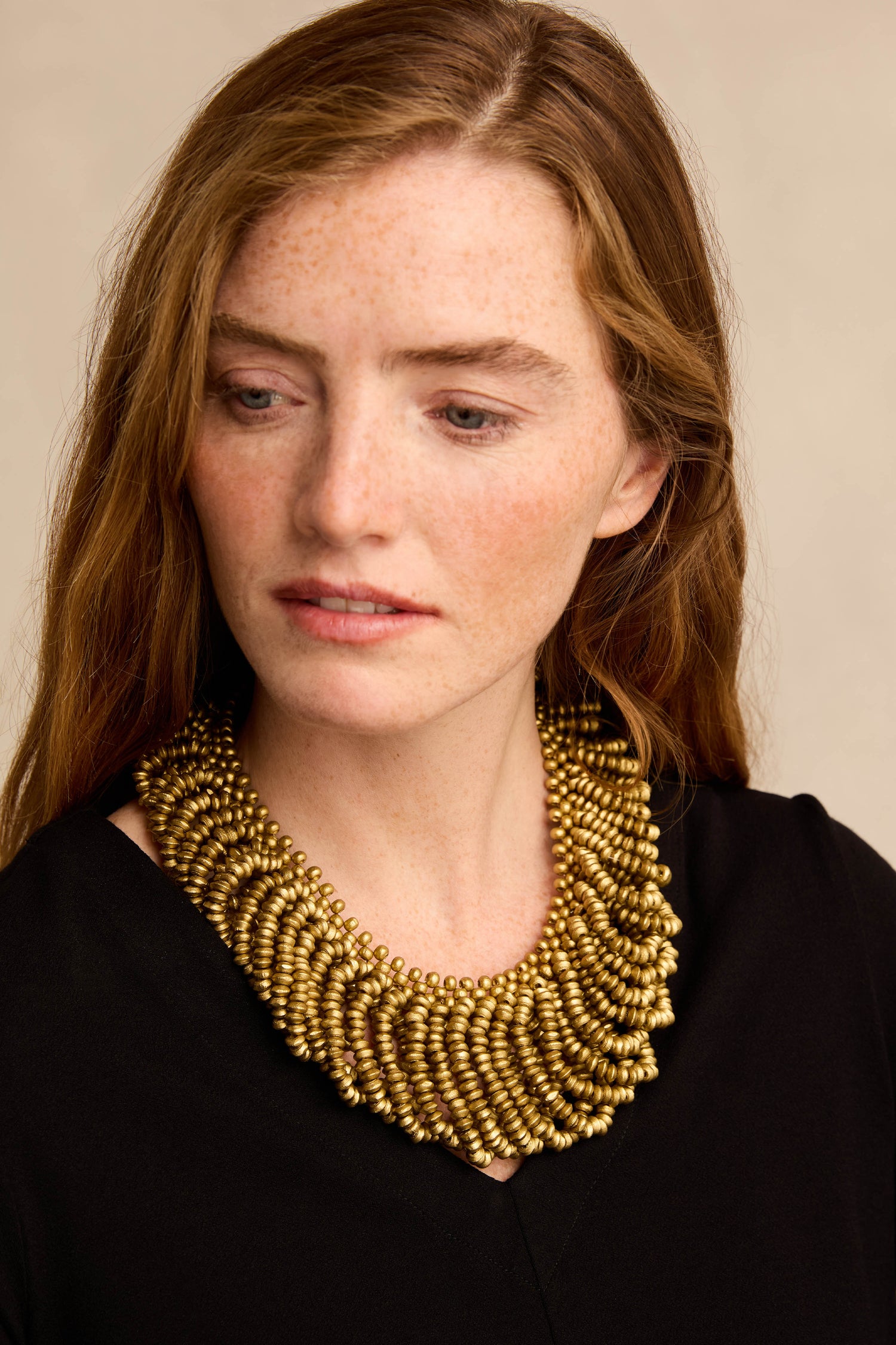 A woman with long brown hair, dressed in a black top and looking slightly to the right against a neutral background, showcases the Gold Beaded Collar Necklace. The layered gold piece adds an elegant touch to her sophisticated look.