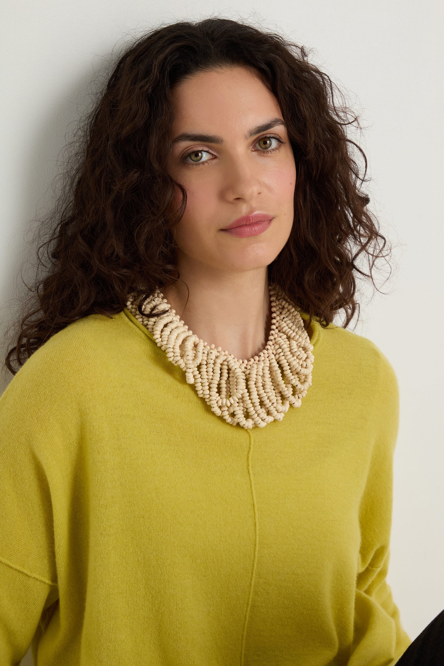 A woman with long curly hair wears a yellow sweater and the Natural Beaded Collar Necklace, standing against a plain light-colored wall.