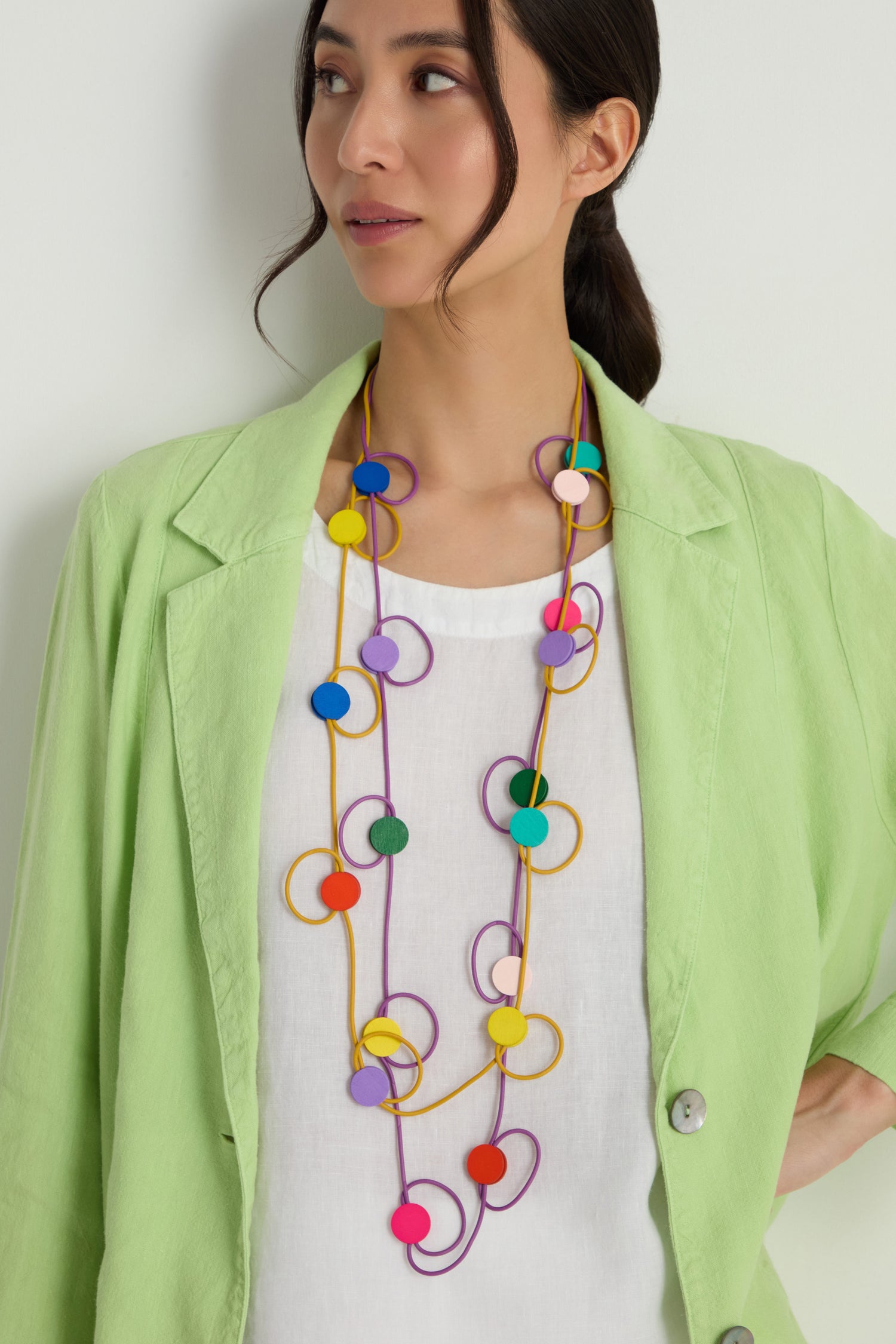 A woman stands against a plain light background, wearing a white top under a light green blazer and accessorizing with the Rainbow Circle Hoop Necklace, a lightweight piece of contemporary jewelry.