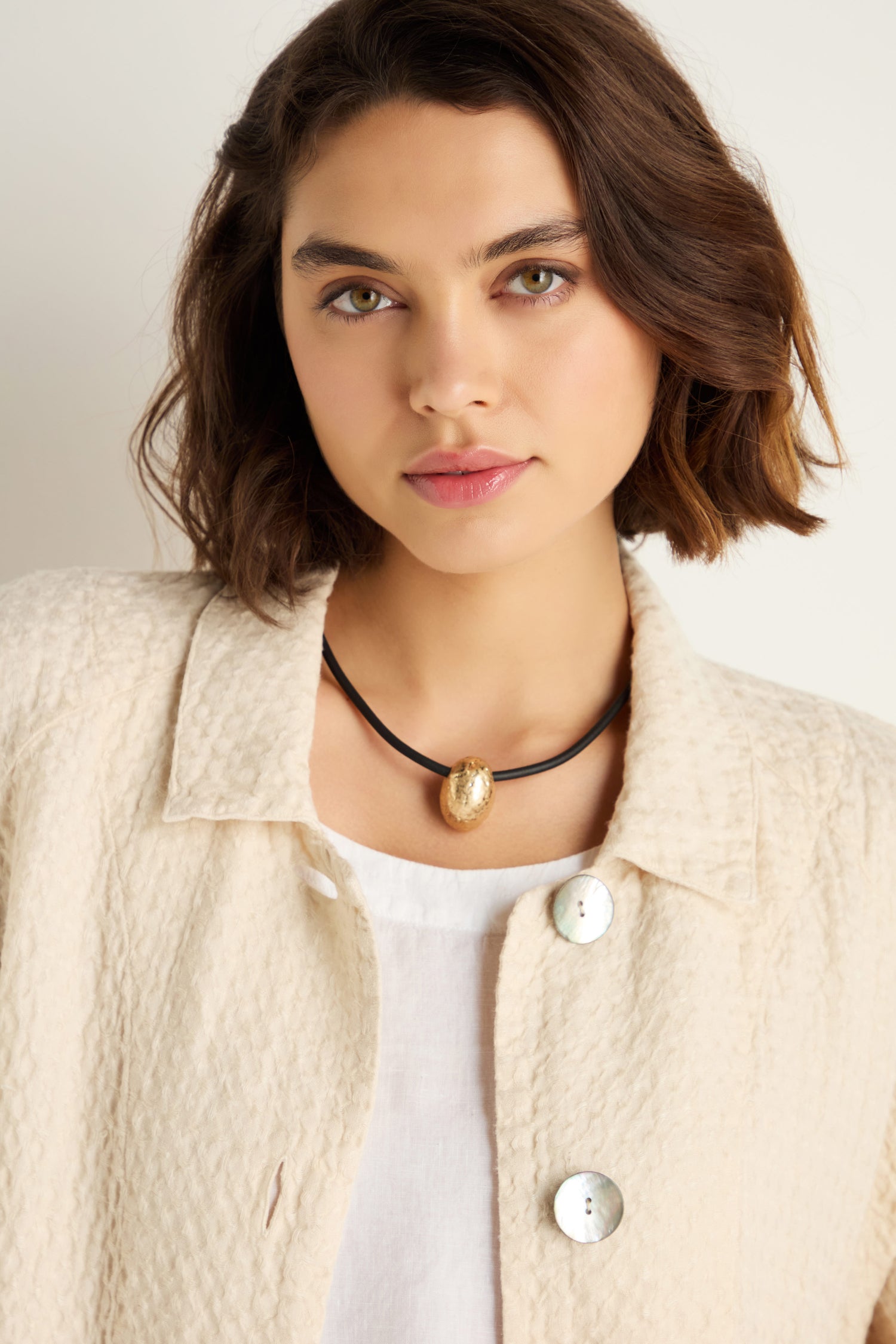 A woman with short brown hair wears a cream textured jacket, white top, and the Hammered Pebble Necklace on a black rubber cord, standing against a light background.