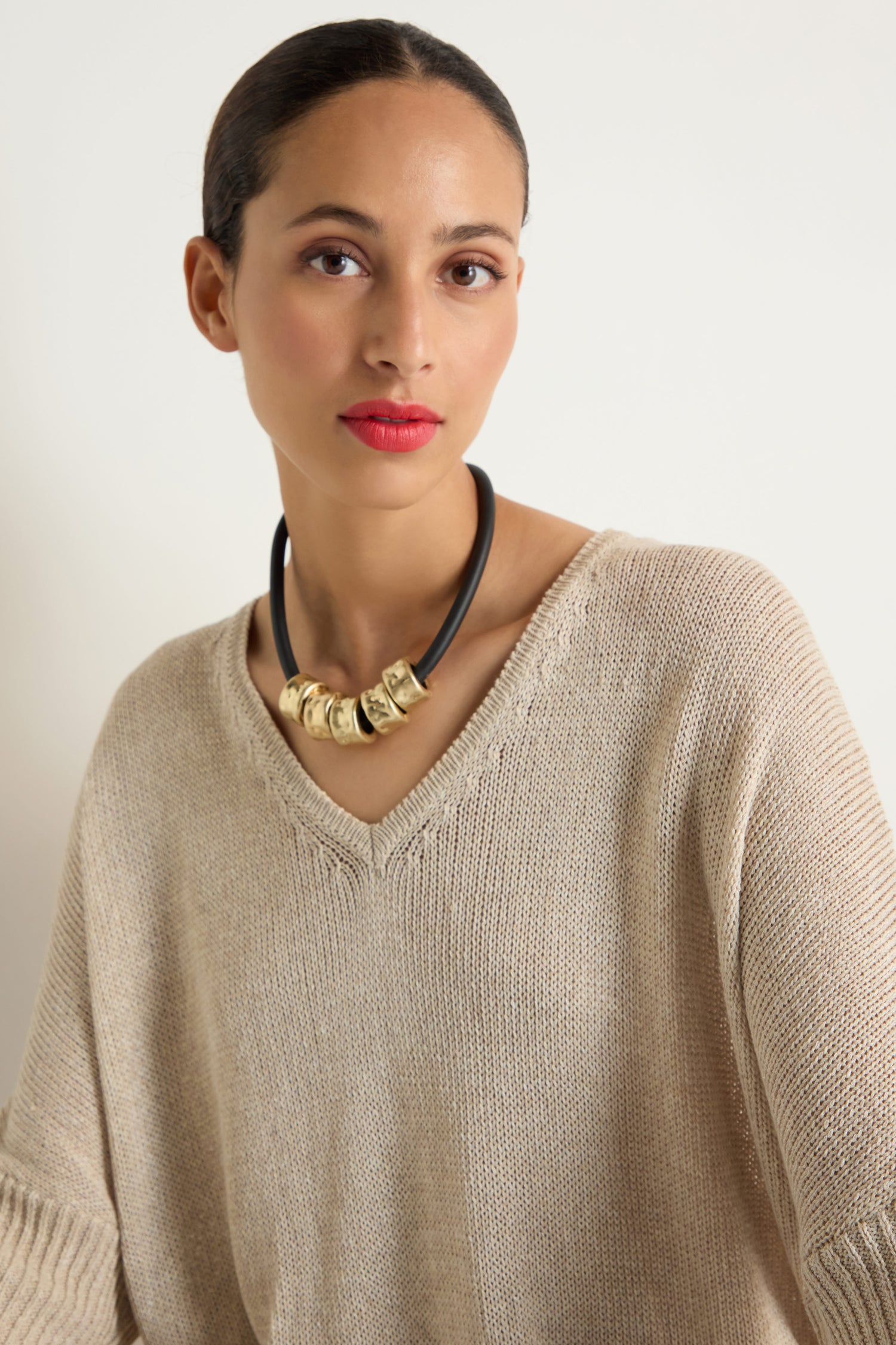 A woman with wavy brown hair in a blue top wears the Hammered Cylinders Necklace, featuring gold beads, as she gazes slightly to the side against a plain background.
