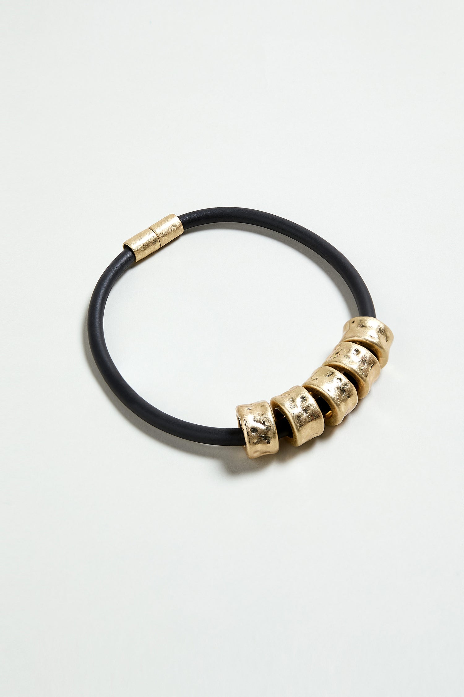 The Hammered Cylinders Necklace features five gold metallic beads and a gold magnetic clasp on a black cord, capturing modern elegance and showcased beautifully against a light background.