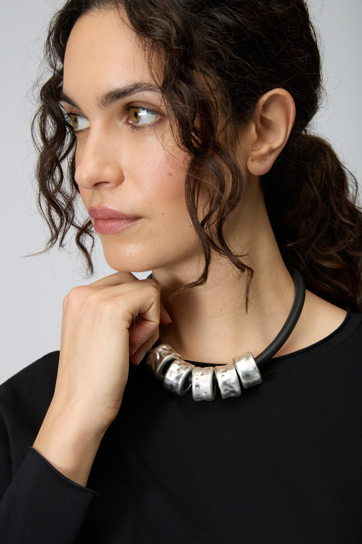 Woman with curly dark hair, wearing a black top and the Hammered Cylinders Necklace, looks to the side with her hand near her chin.