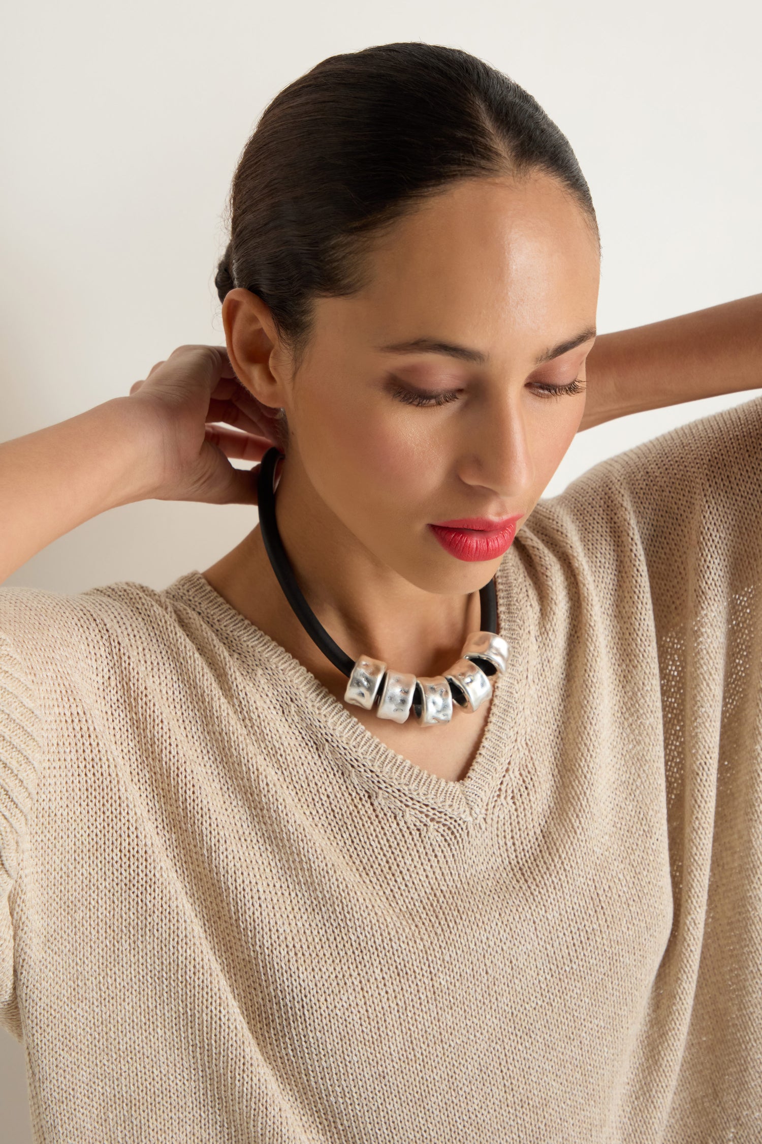 Woman with curly dark hair, wearing a black top and the Hammered Cylinders Necklace, looks to the side with her hand near her chin.