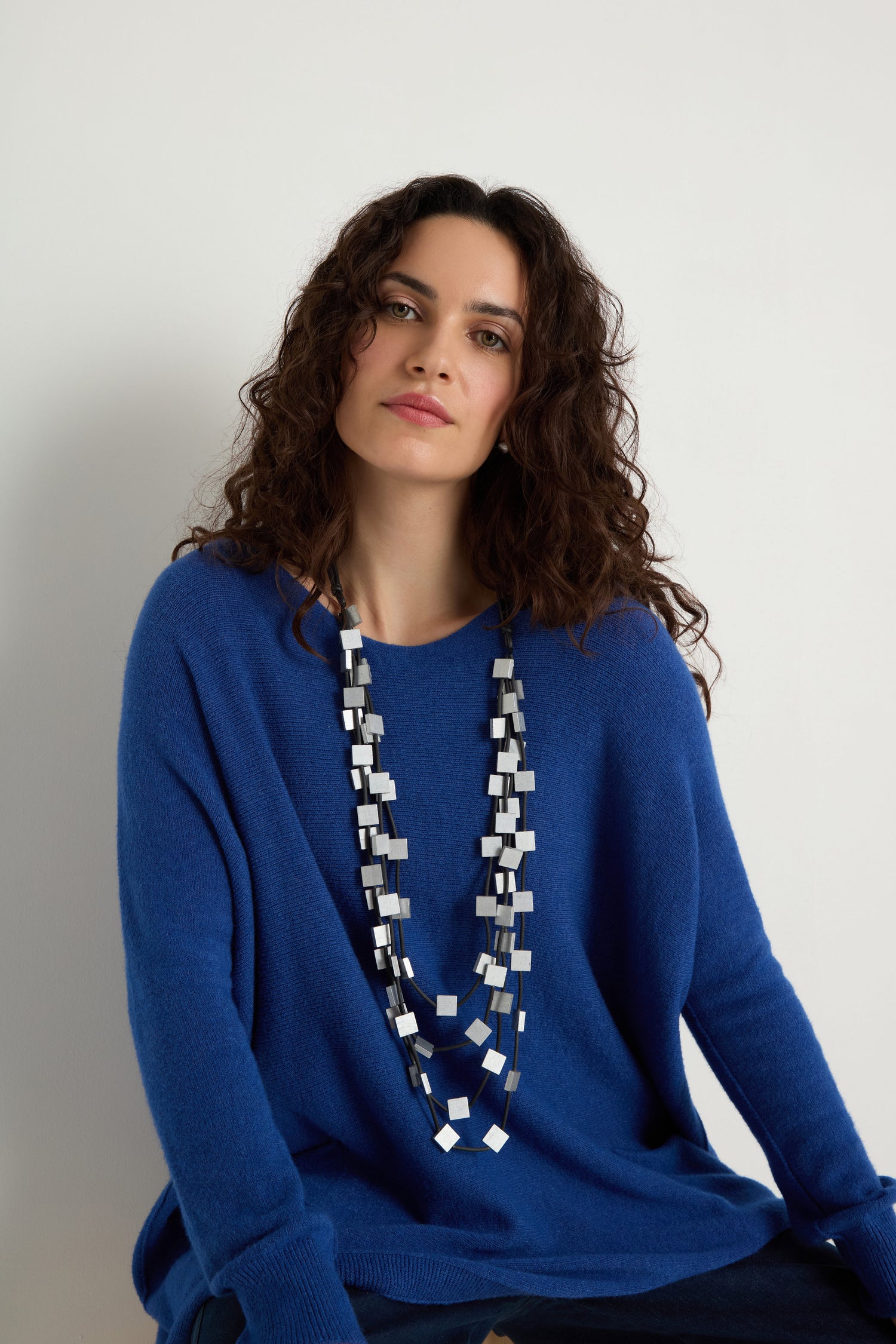 A woman with wavy brown hair wears a blue sweater and the Cascade Squares Necklace, featuring multi-strand geometric black, white, and silver-tone squares, seated against a plain white background.