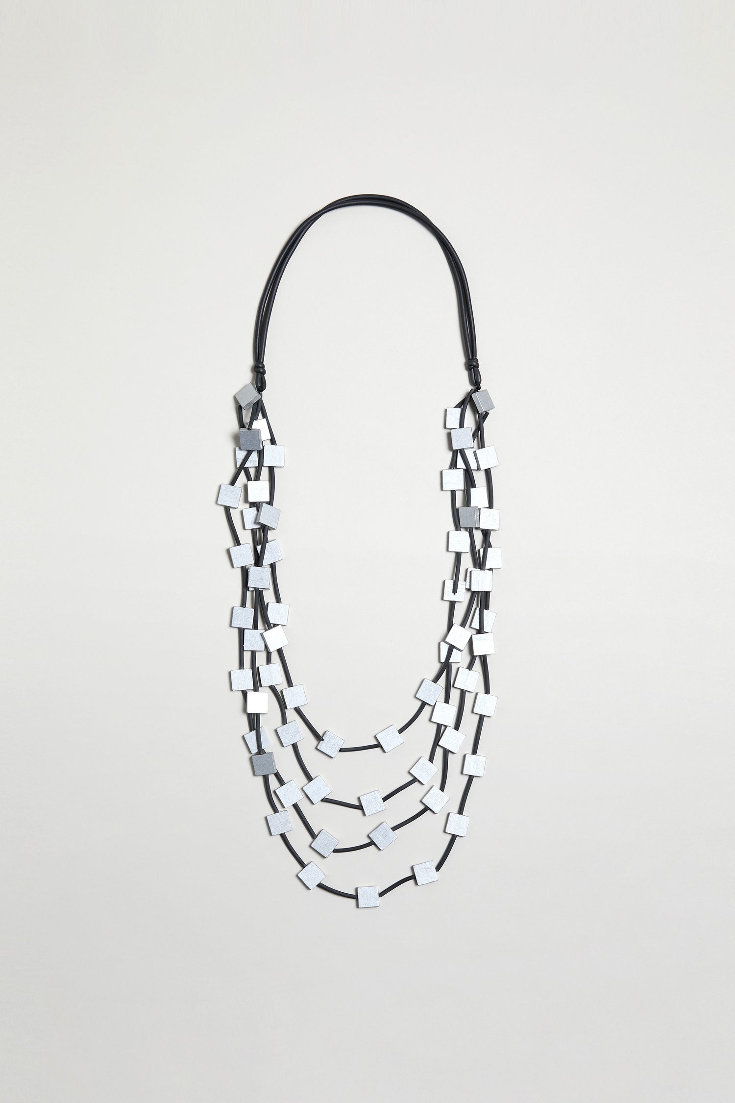 The Cascade Squares Necklace features rectangular silver-tone squares on a black cord, displayed against a plain light background.