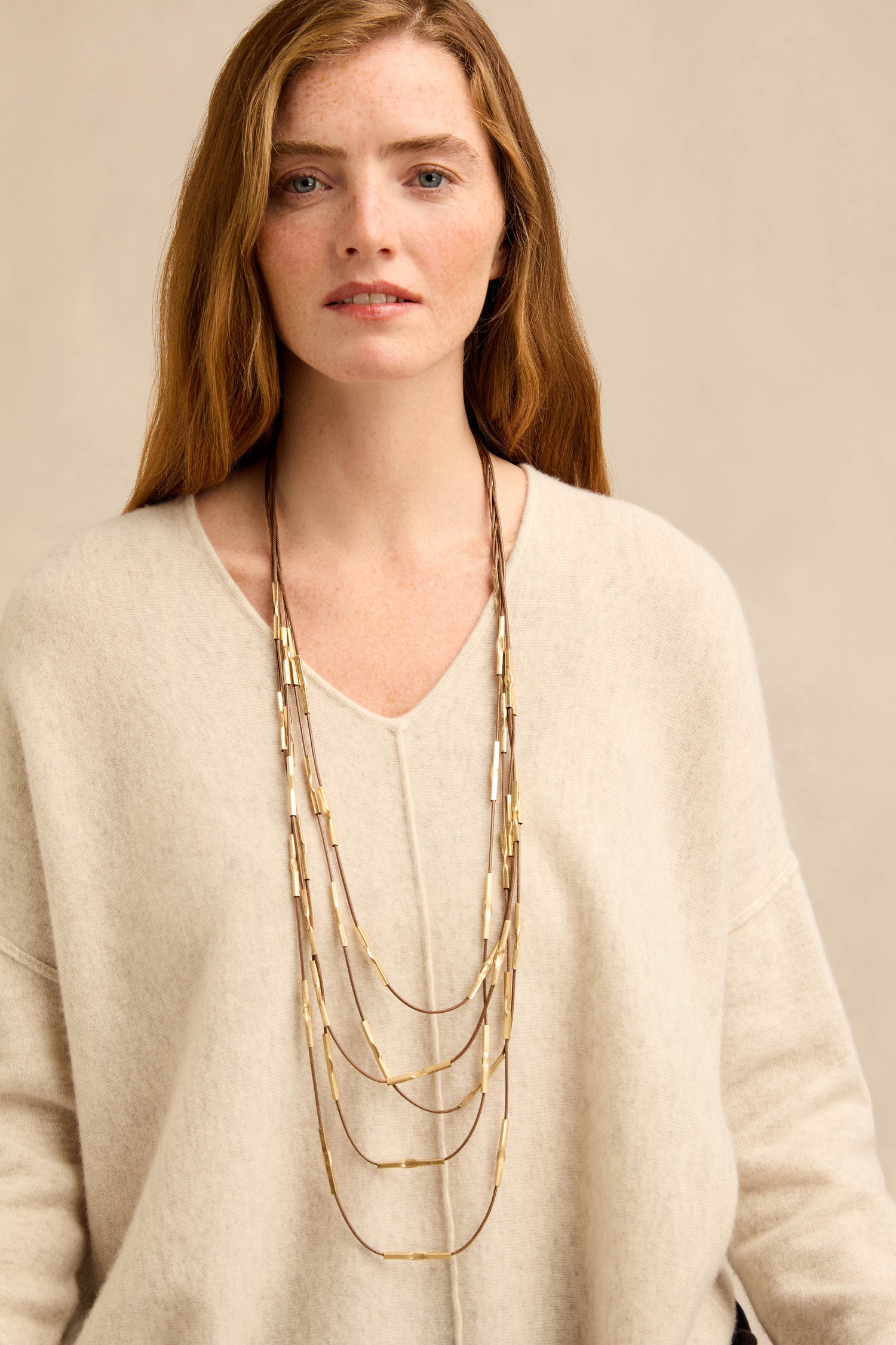 A woman with long auburn hair wears a cream sweater and the Leather and Bead Cascade Necklace, standing against a plain beige background.