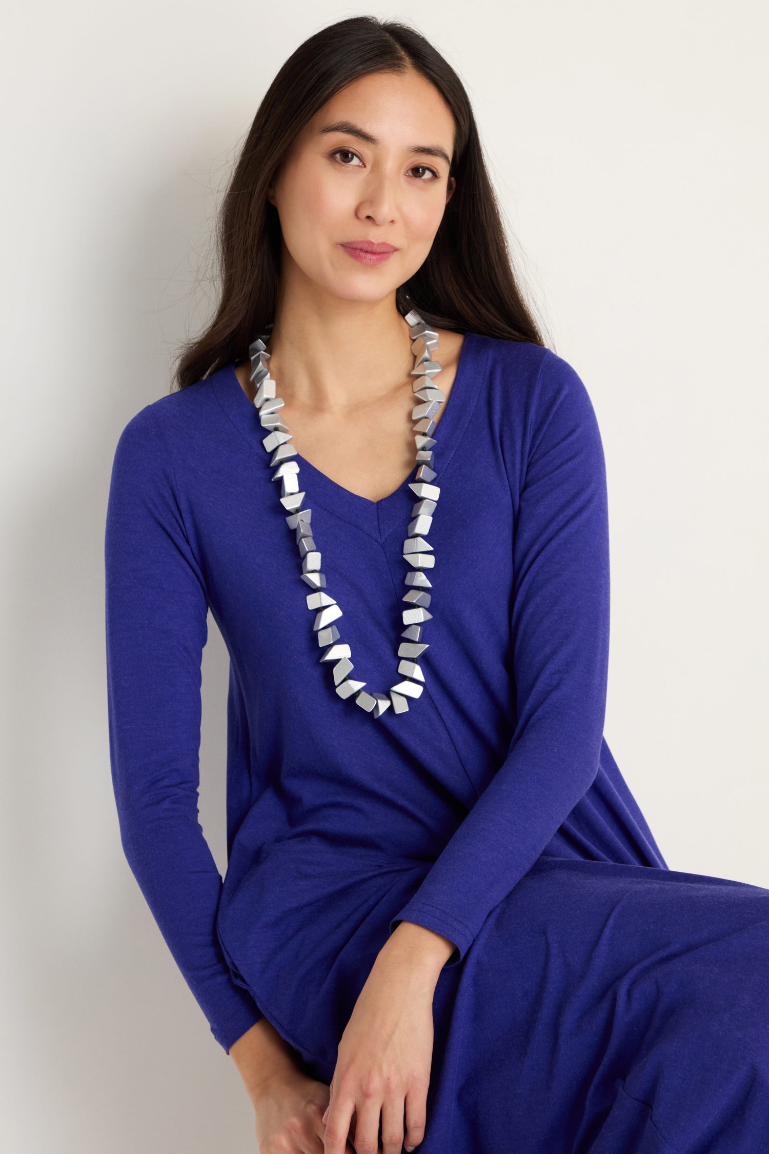 Woman with long dark hair wearing a blue long-sleeve dress and the Long Wooden Metallic Necklace, seated against a plain light background.