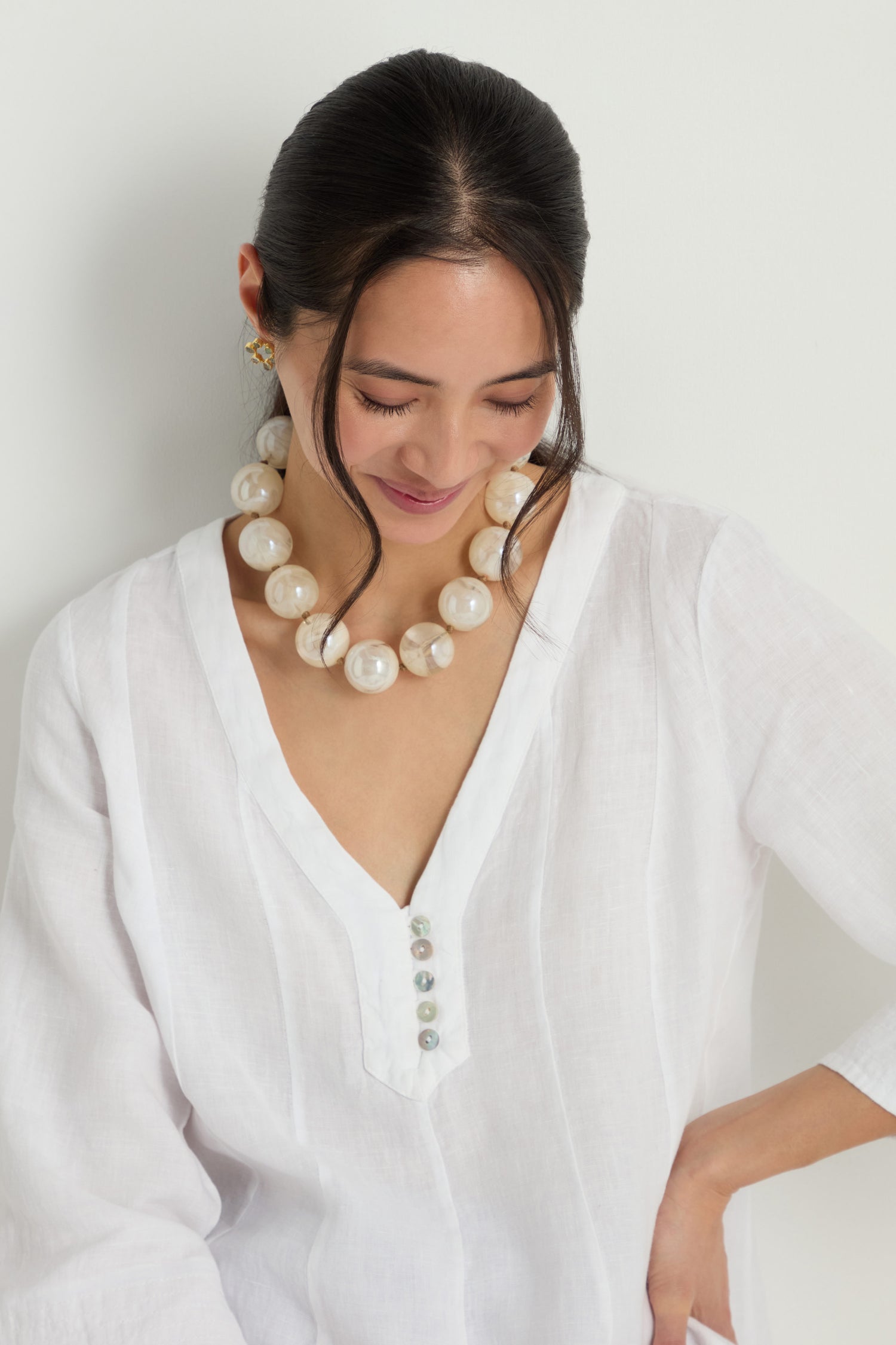 A woman in a white v-neck blouse wears the Marble Spheres Necklace as she looks down and smiles, standing against a plain white background.