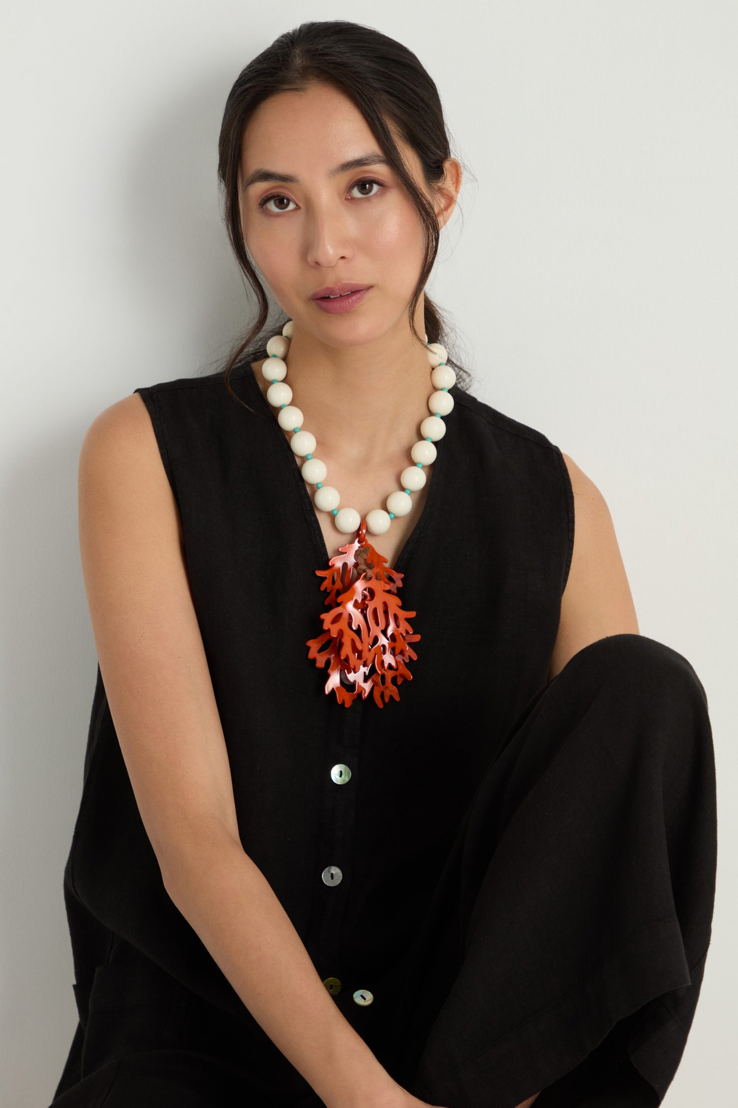 A woman in a black sleeveless outfit sits with one knee up, wearing the Coral Resin Short Pendant Necklace featuring a bold red coral pendant.