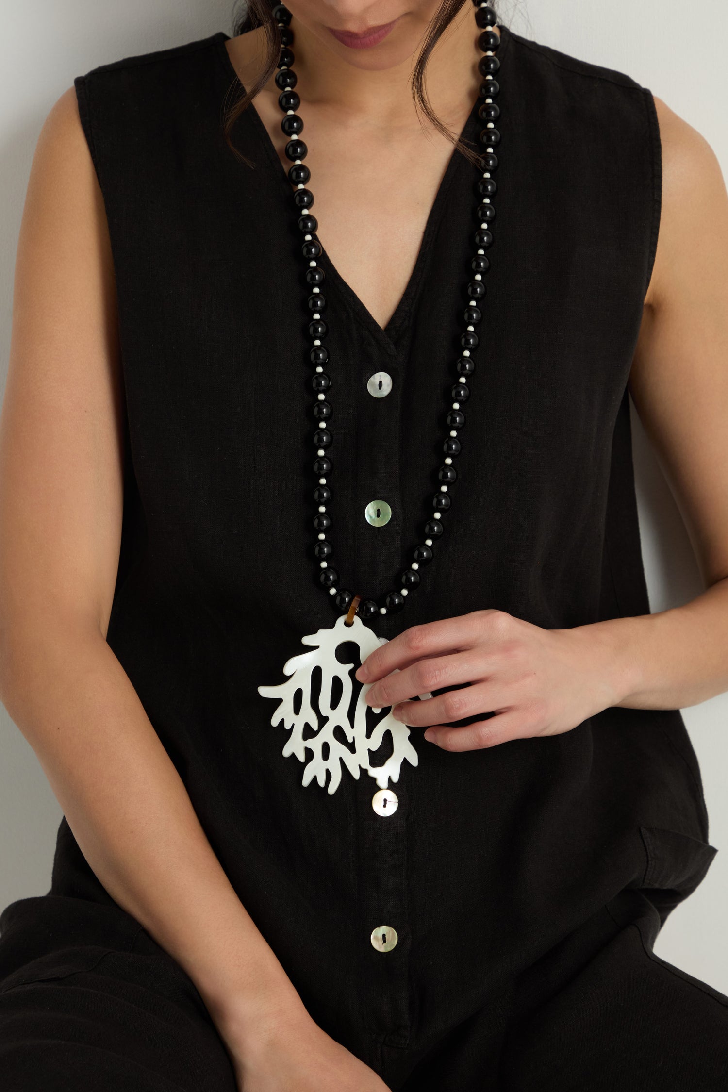 A person wears a sleeveless black button-up top and the Coral Resin Pendant Necklace with black beads, holding its large, sculptural coral resin pendant in one hand.