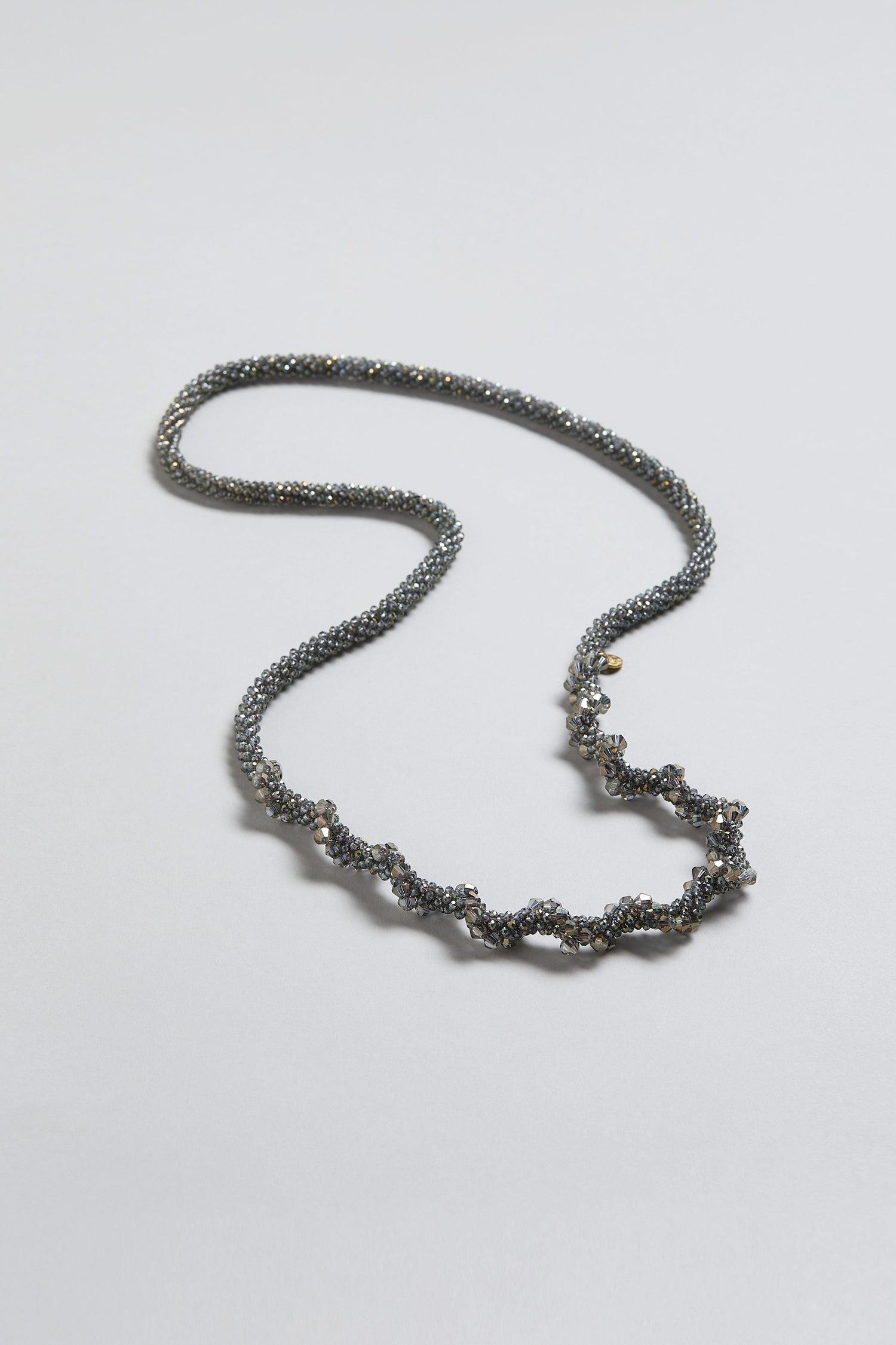 The Glacier Spiral Necklace features a twisted beadwork design with metallic and dark-colored beads, presented on a simple light background.