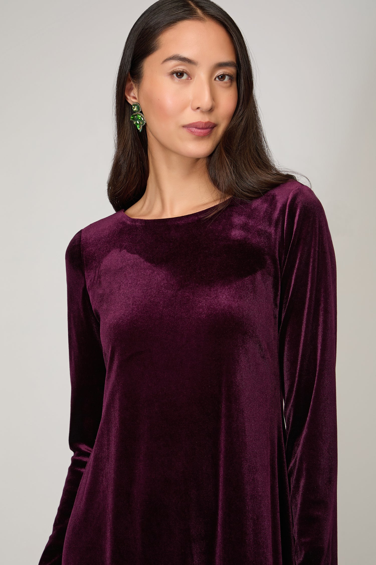Woman with long dark hair wears the Velvet Jersey Maxi Dress in deep purple paired with green earrings, standing against a plain light background for an elegant occasionwear look.