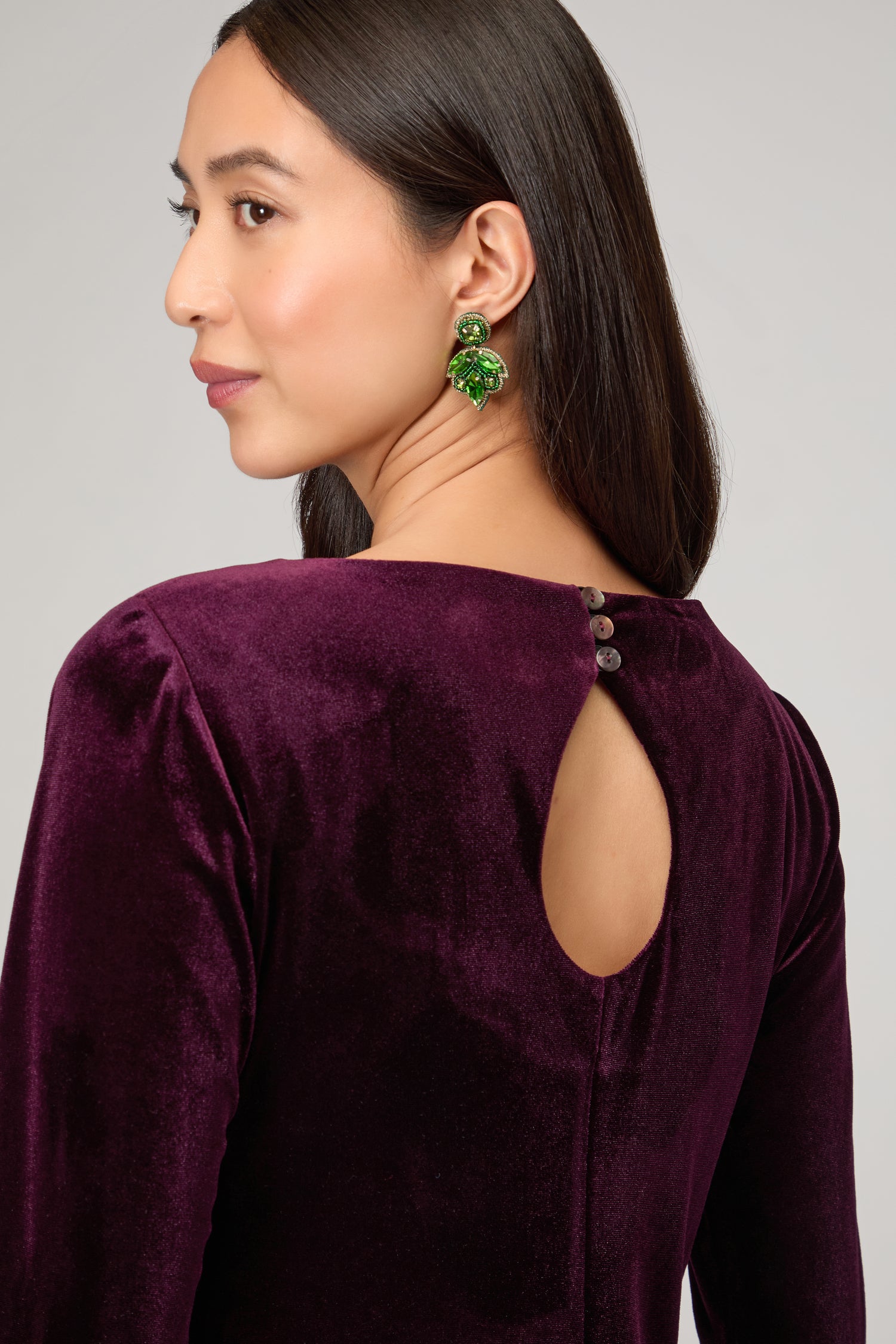 A woman poses confidently in the Velvet Jersey Maxi Dress, deep purple with a keyhole back and button details, styled with green statement earrings—the perfect choice for striking occasionwear.