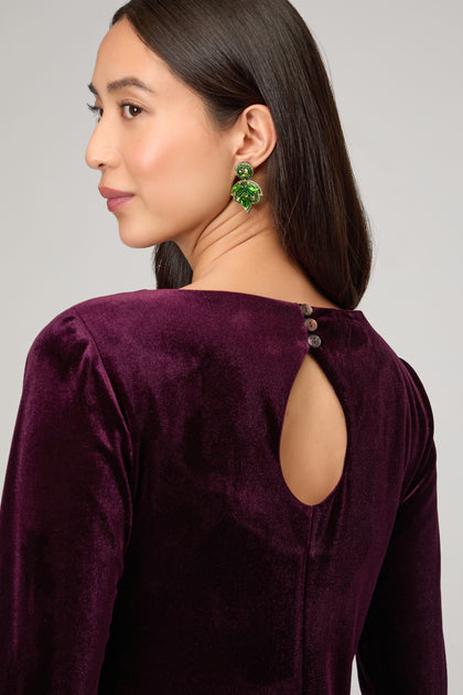 A woman poses confidently in the Velvet Jersey Maxi Dress, deep purple with a keyhole back and button details, styled with green statement earrings—the perfect choice for striking occasionwear.