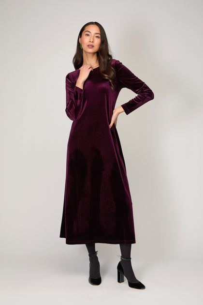 A woman stands against a plain background wearing the Velvet Jersey Maxi Dress with long sleeves and ankle-length dark purple velvet, paired with black heeled shoes—an elegant occasionwear choice.