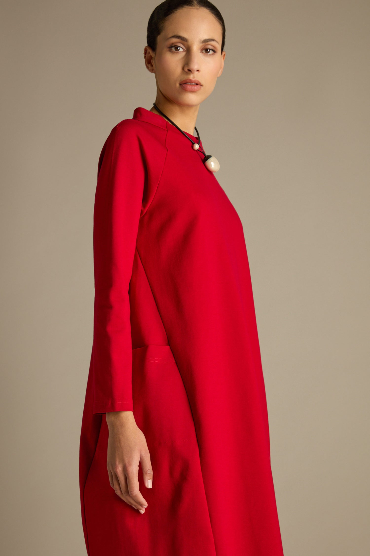 A person is elegantly posed against a plain background, wearing a red Ponte Pocket Dress characterized by long sleeves, raglan armholes, and a high neckline, complemented by a pearl necklace.