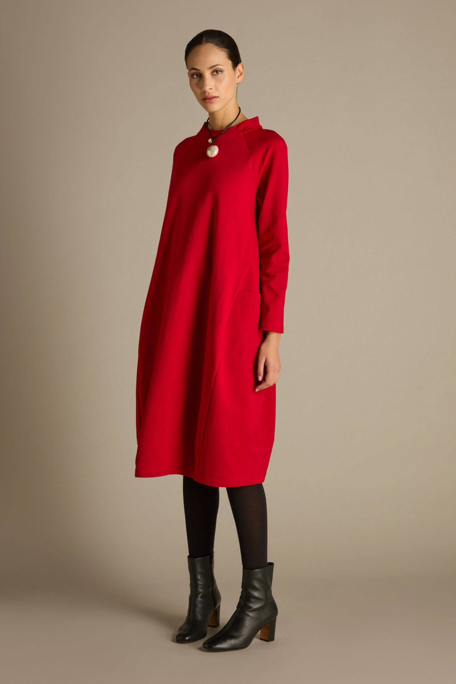 A person dressed in a Ponte Pocket Dress in a vibrant red hue, complemented by black tights and ankle boots, poses gracefully against a simple backdrop.