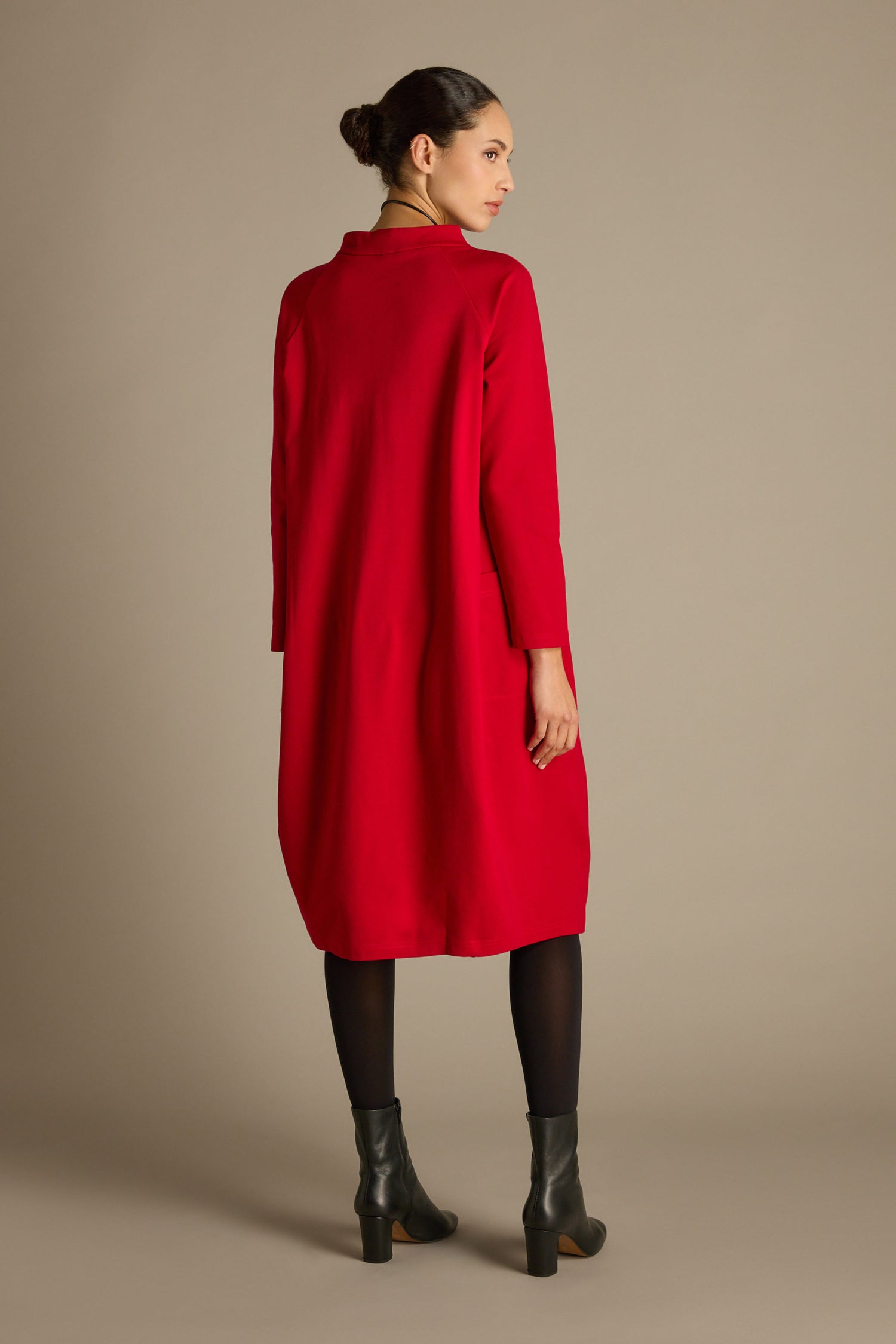A woman stands with her back to the camera, wearing a long red Ponte Pocket Dress of calf-grazing length, paired with black leggings and ankle boots. Her hair is tied back in a neat bun, enhancing the chic cocoon shape silhouette of her ensemble.