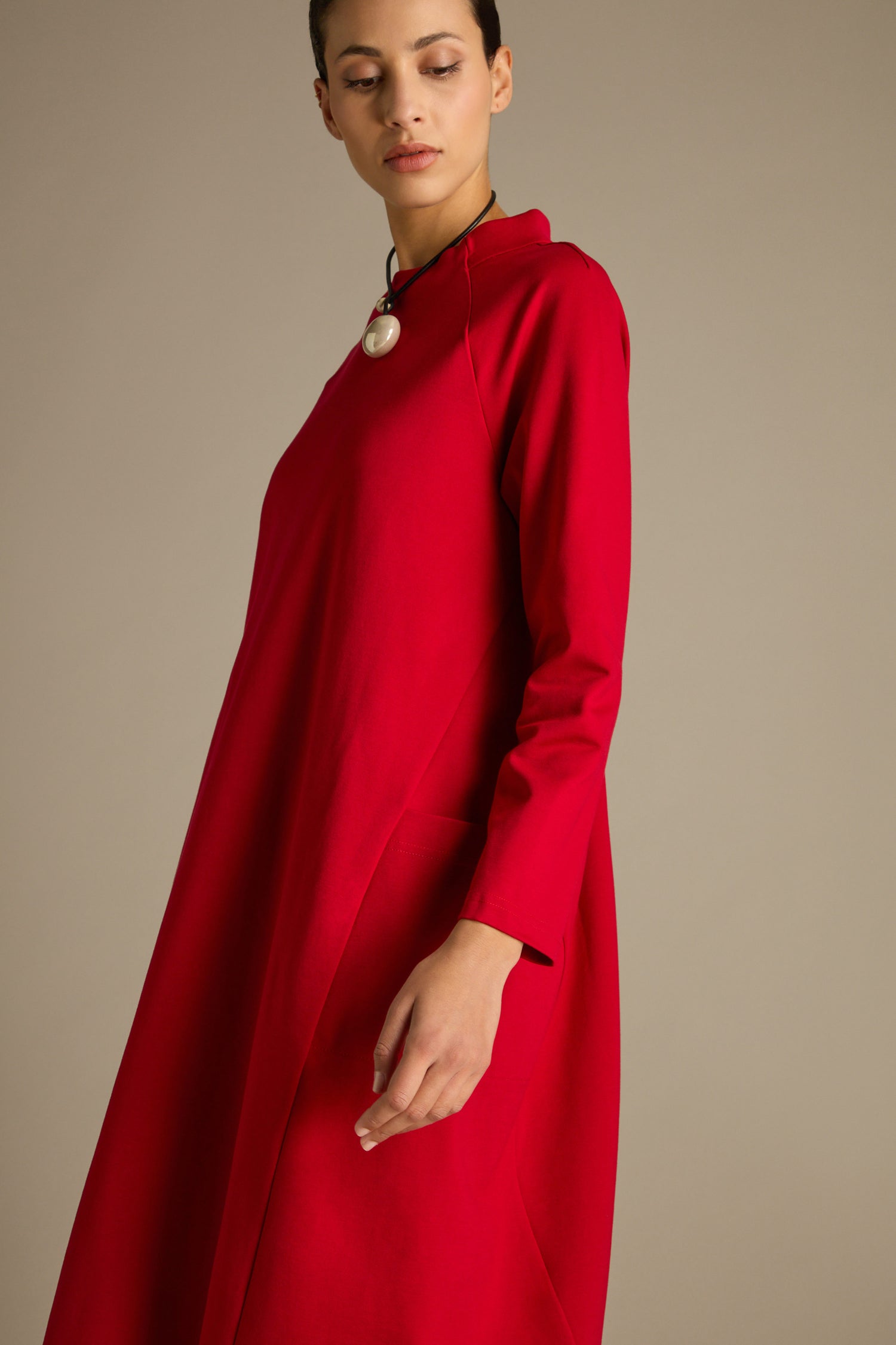 The person is elegantly attired in the form-fitting, calf-grazing length Ponte Pocket Dress featuring long sleeves and complemented by a black necklace, standing against a neutral backdrop.
