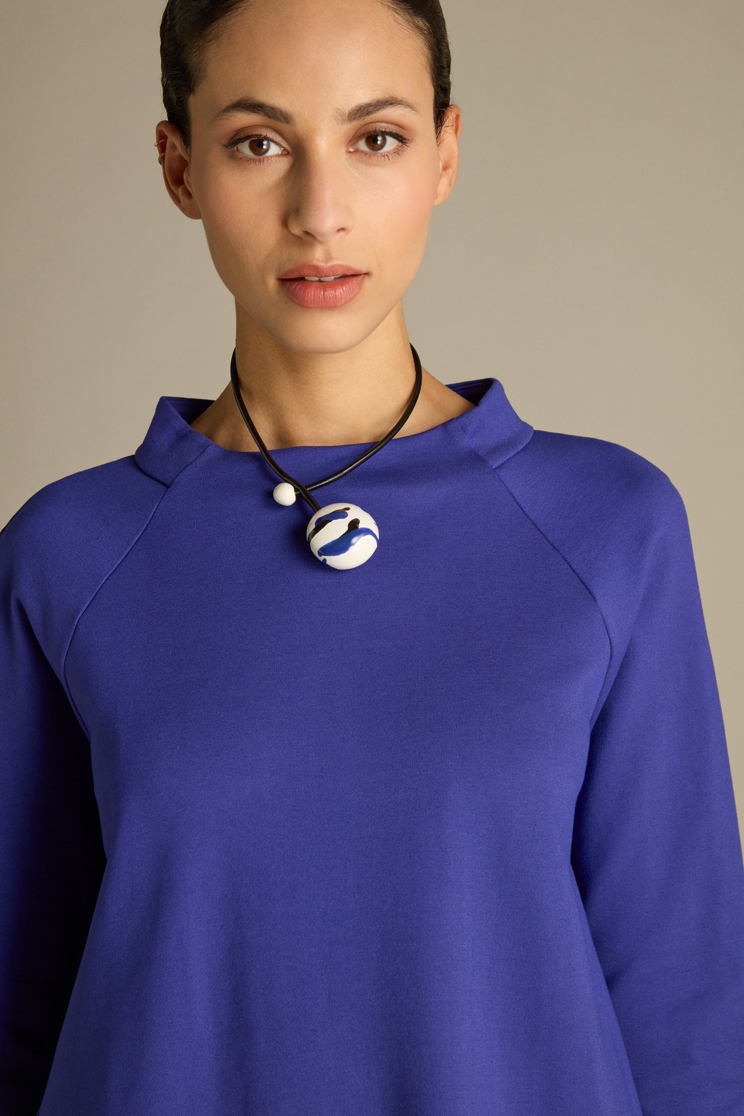 A person wearing a blue Ponte Pocket Dress featuring raglan armholes and a ceramic pendant necklace stands against a neutral background.