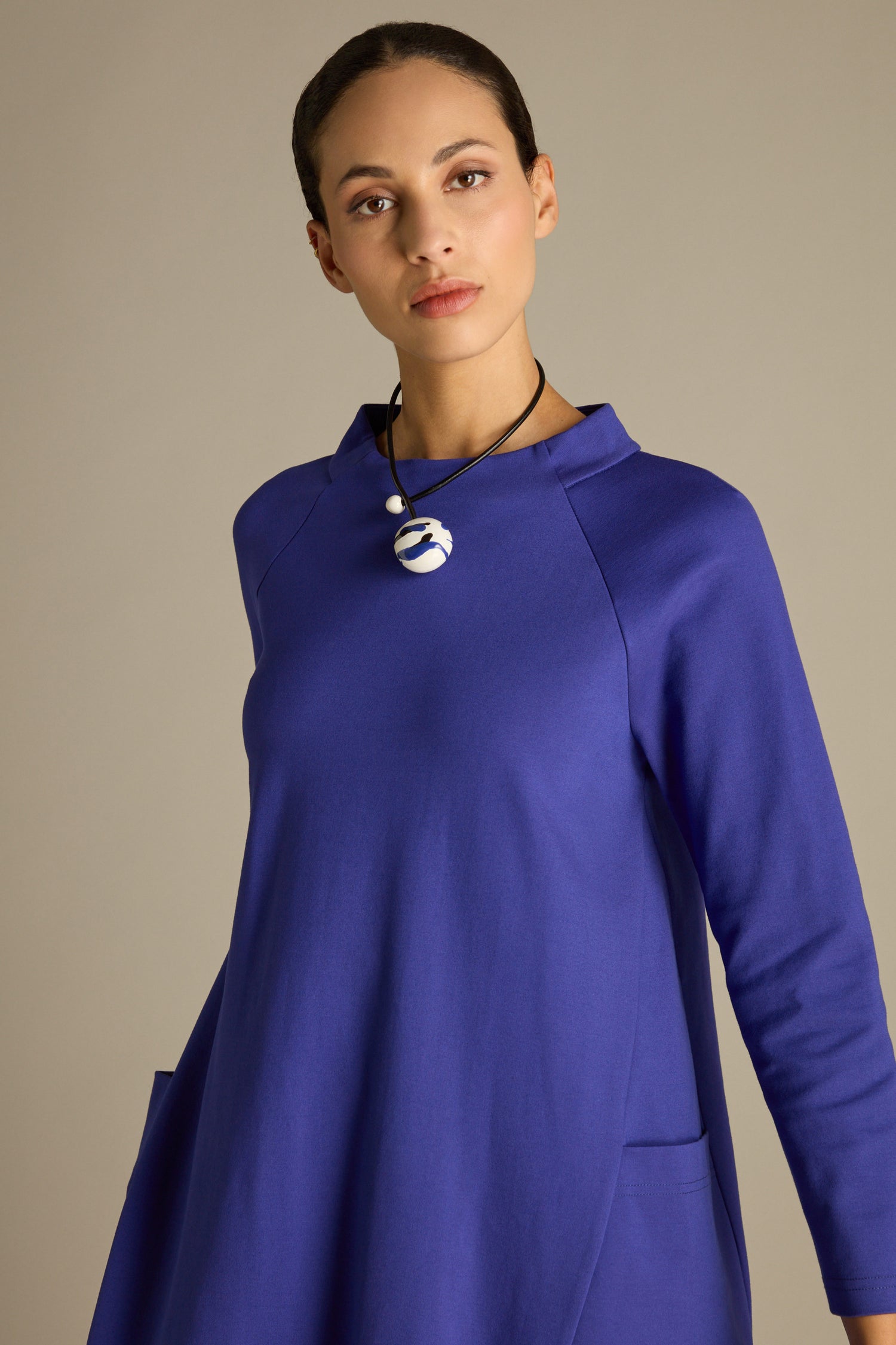 A person wearing an elegant cocoon-shaped dress named the Ponte Pocket Dress in blue, paired with a black and white pendant, stands against a plain background.
