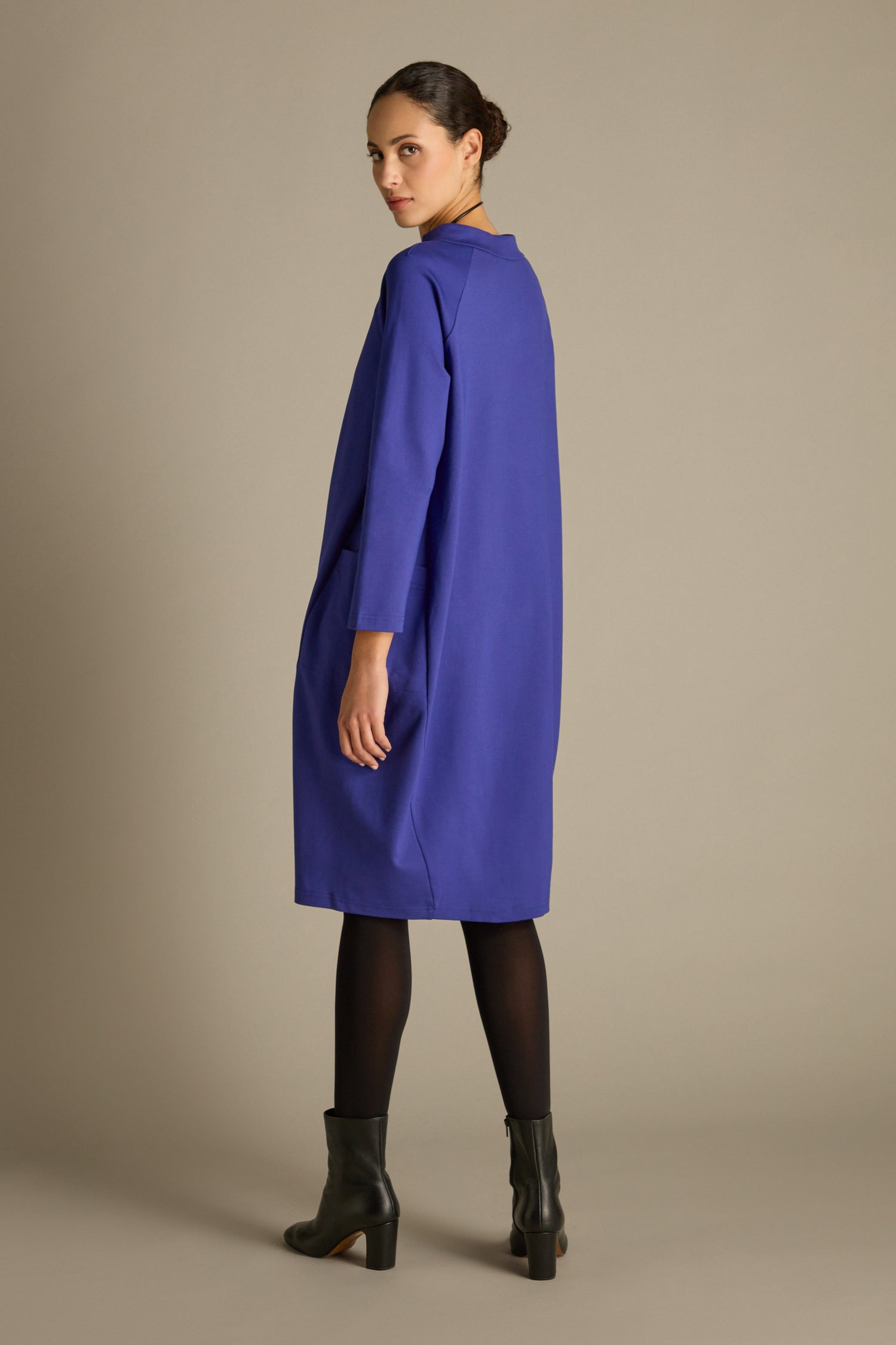 A woman wearing a long-sleeved Ponte Pocket Dress in royal blue paired with black tights and ankle boots stands against a beige backdrop, the dress's cocoon shape cascading gracefully as she gazes over her shoulder.