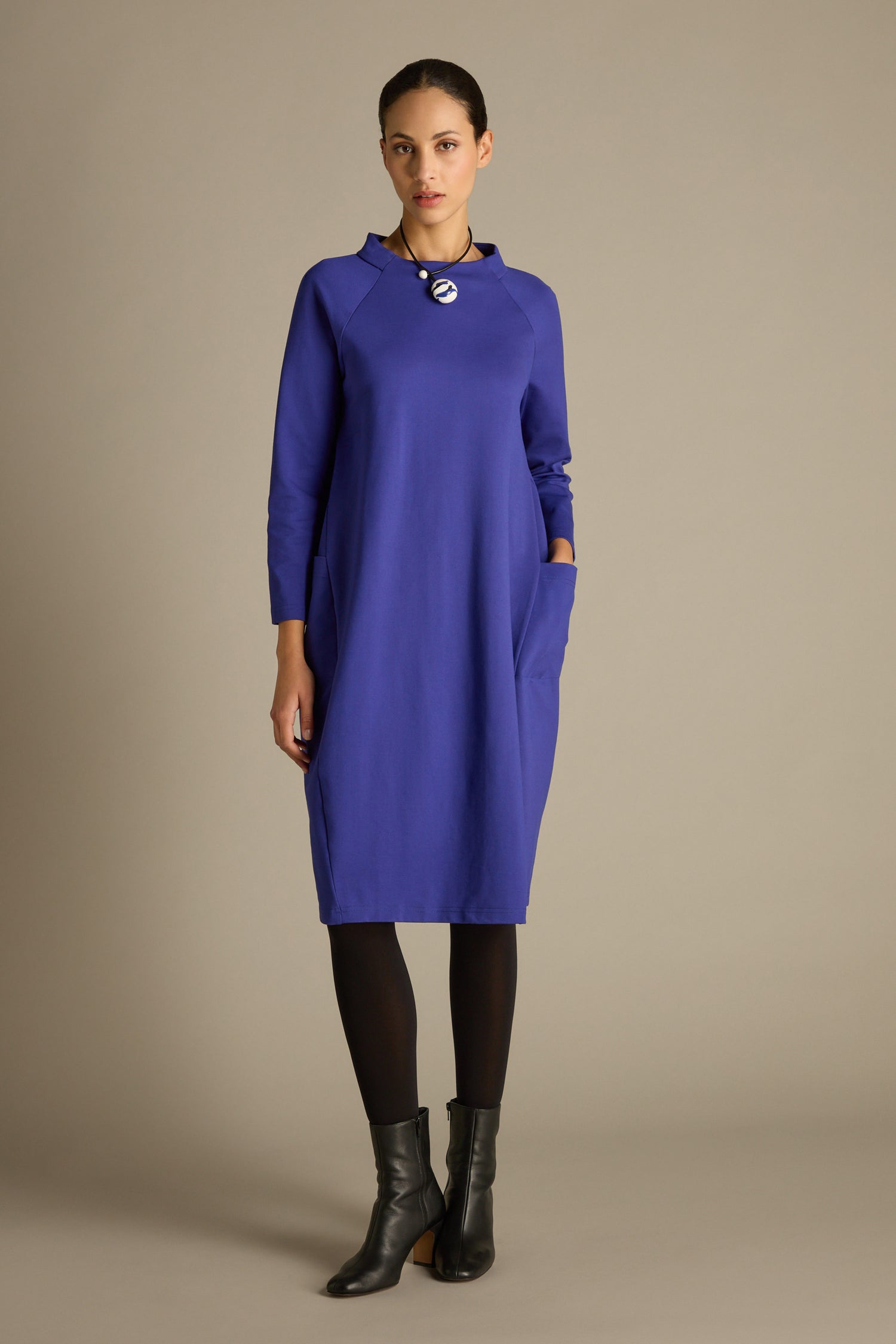 A person stands in a blue Ponte Pocket Dress, featuring raglan-style armholes, paired with black leggings and ankle boots, against a plain background.