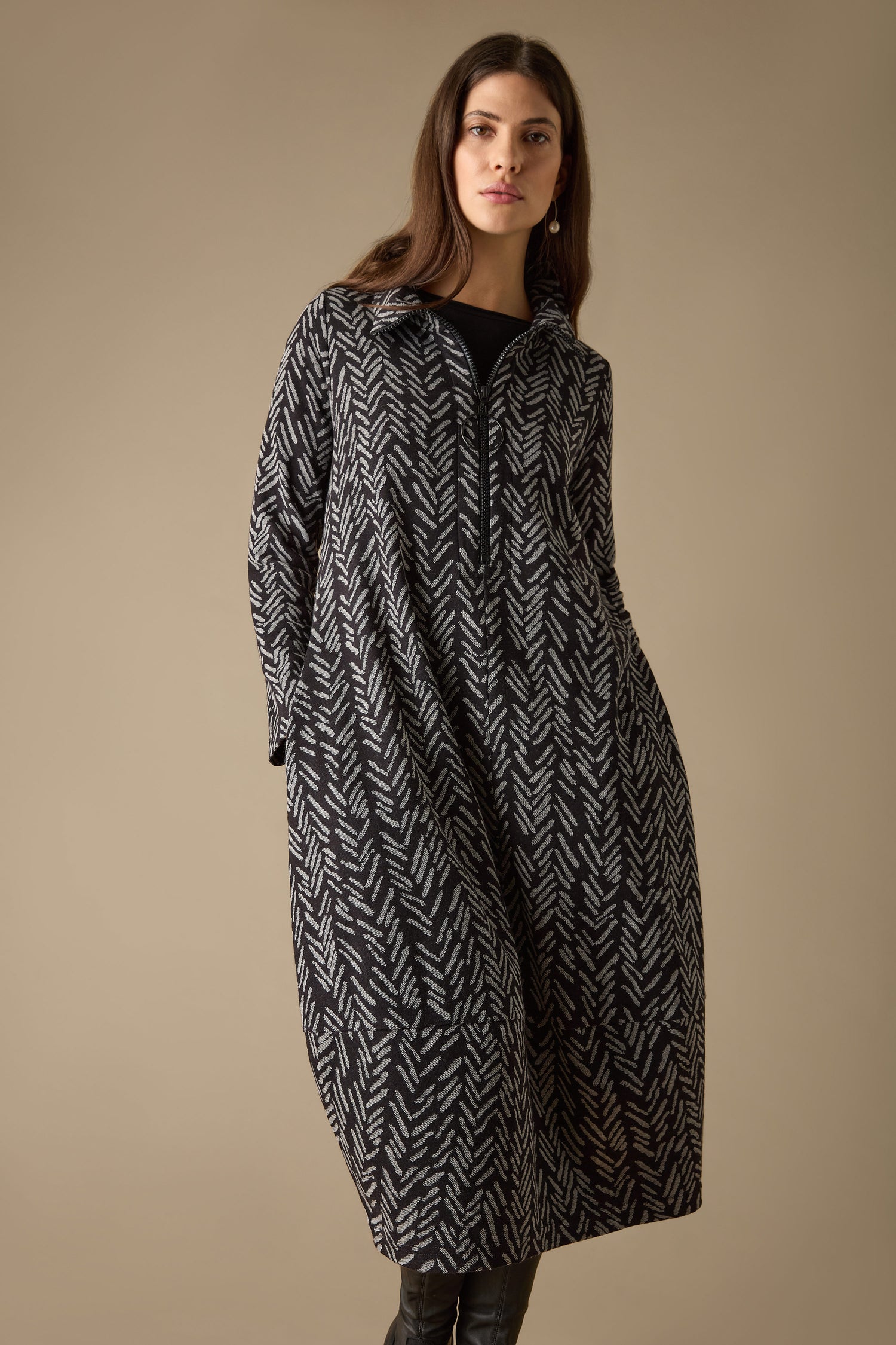 Against a simple backdrop, an individual poses elegantly in a Herringbone Jersey Jacquard Dress with patterned long sleeves, where the sophisticated jacquard fabric introduces texture and richness to the chic outfit.