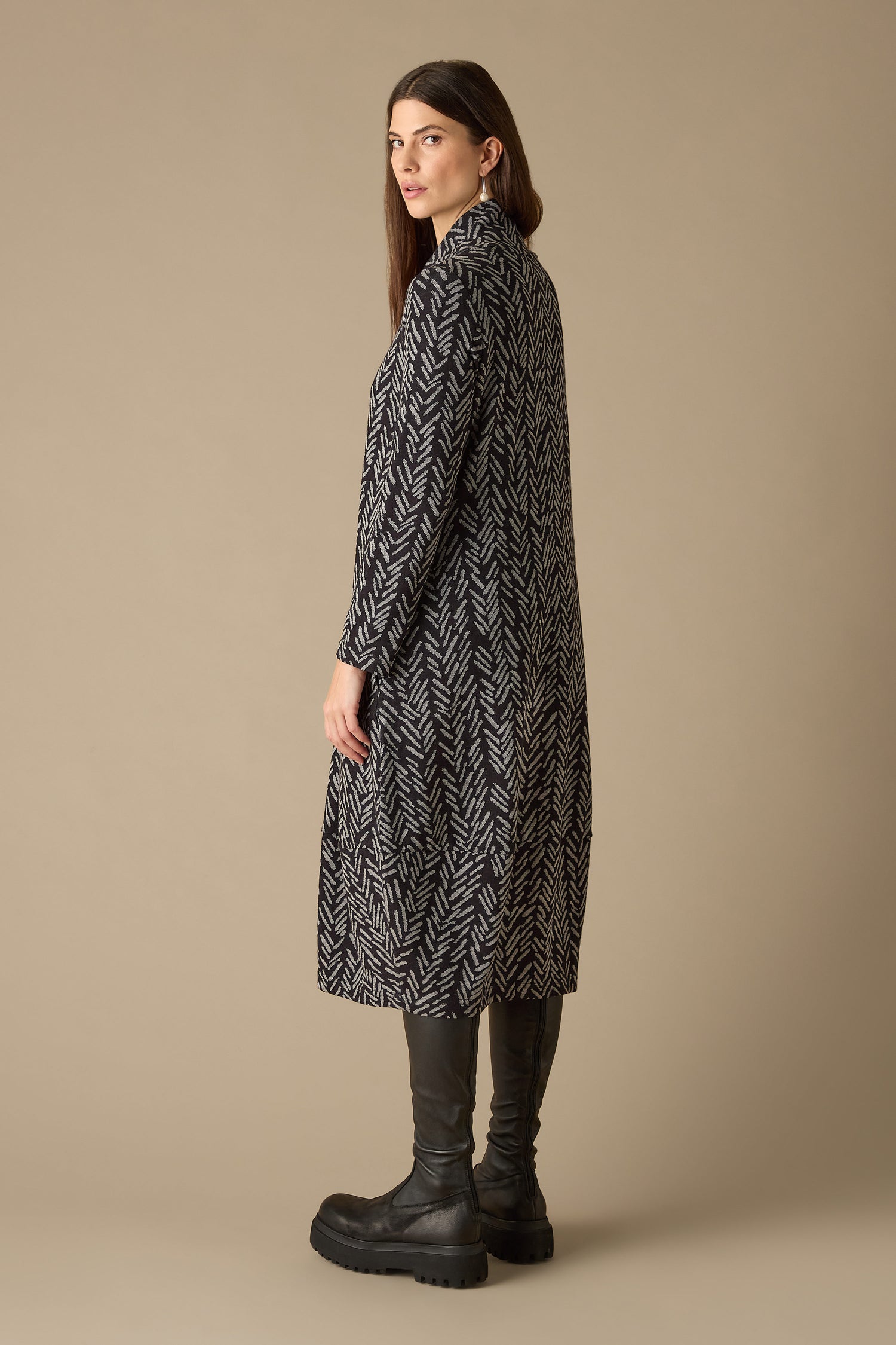A woman stands sideways, wrapped in a Herringbone Jersey Jacquard Dress, paired with black leggings and chunky black boots, against a neutral background.