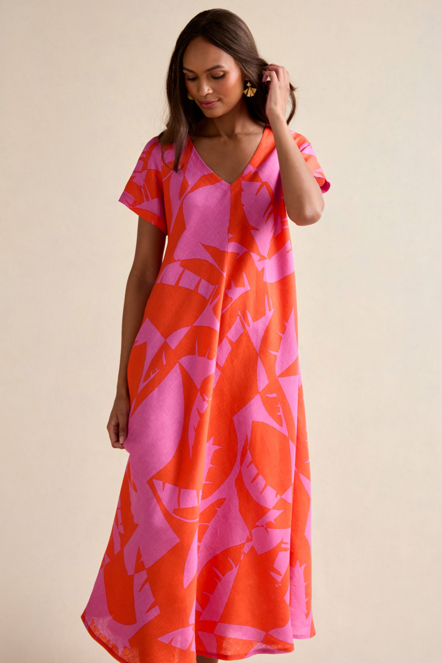 A woman wears the Palm Mirage Linen Flared Dress, featuring a flowing, short-sleeved design with a red and pink abstract leaf pattern, standing against a neutral background.