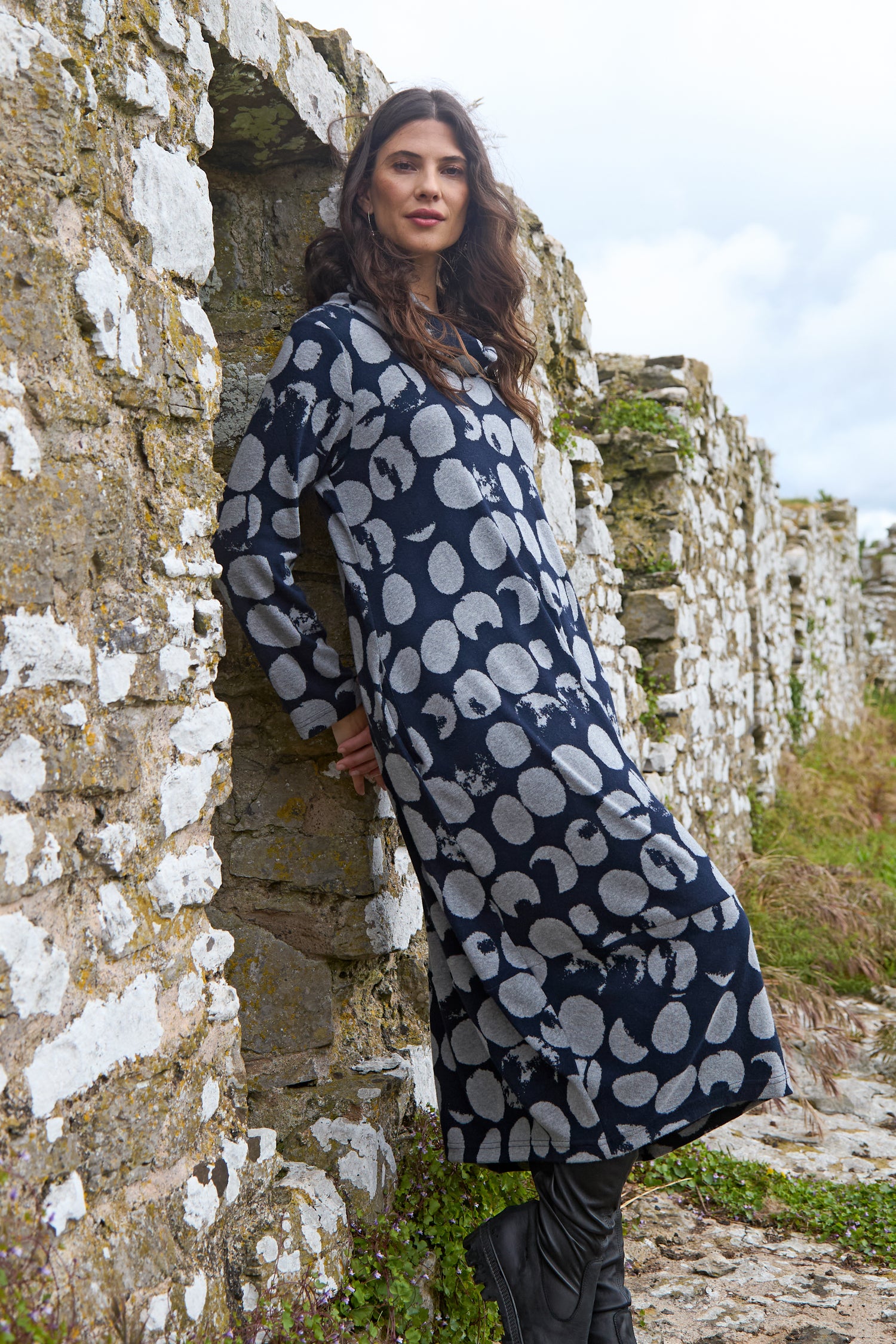 A woman in a long Brushed Jersey Spotted Bubble Dress with a white circular pattern stands against a weathered stone wall outdoors.