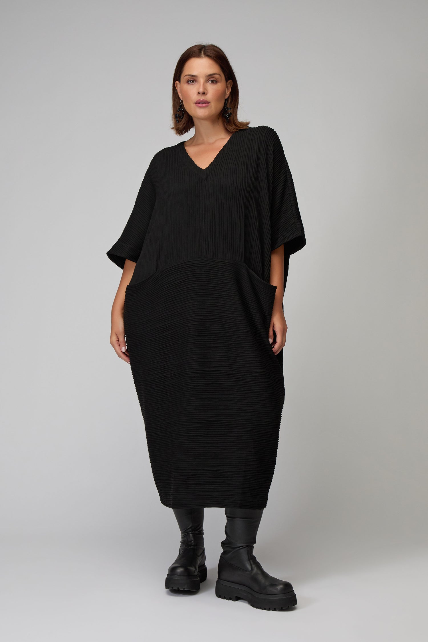 A woman stands in a studio wearing the Textured Wave Jersey Dress, featuring an oversized fit and V-neckline, paired with black chunky boots, against a plain gray background.