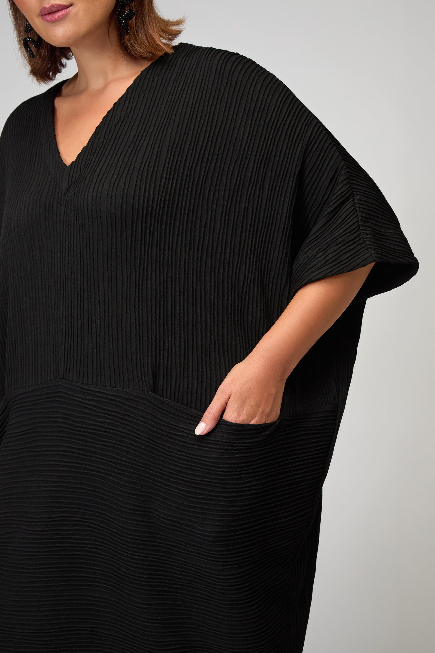 Model wears an oversized fit, black Textured Wave Jersey Dress with V-neckline and short sleeves, shown from shoulders to above the knee. One hand is placed in a side pocket.