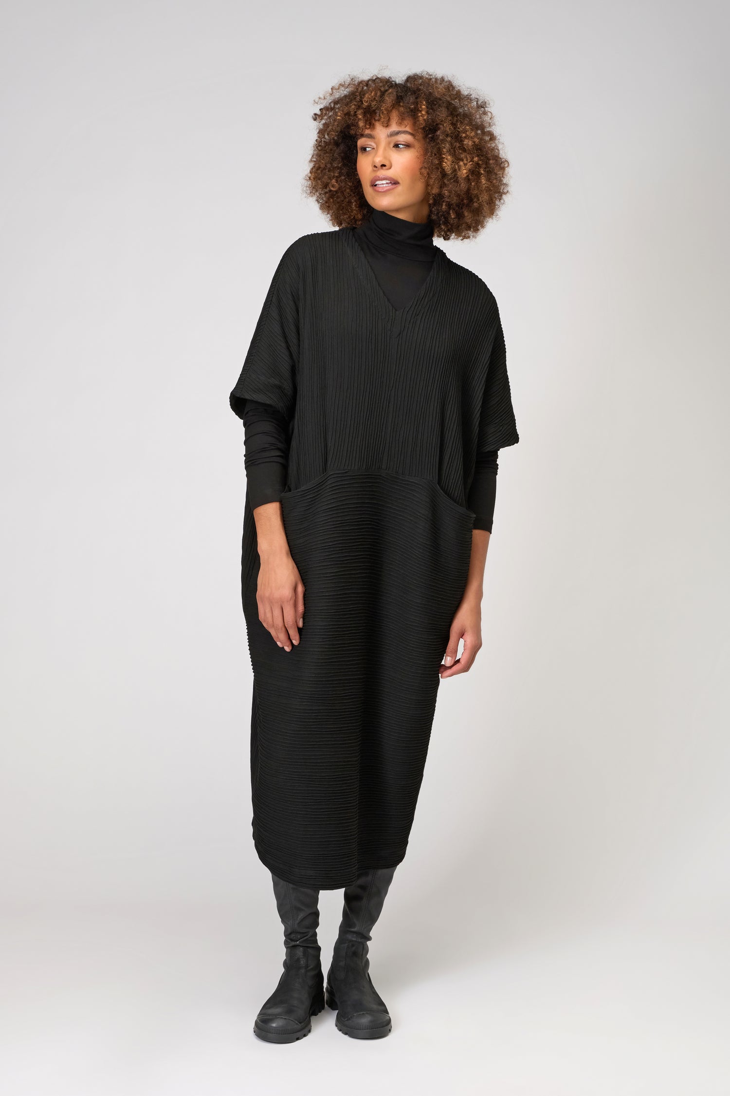 A person wears the Textured Wave Jersey Dress, a loose black kaftan-inspired dress in pleat-effect viscose jersey, over a black turtleneck and boots, standing against a plain light background.