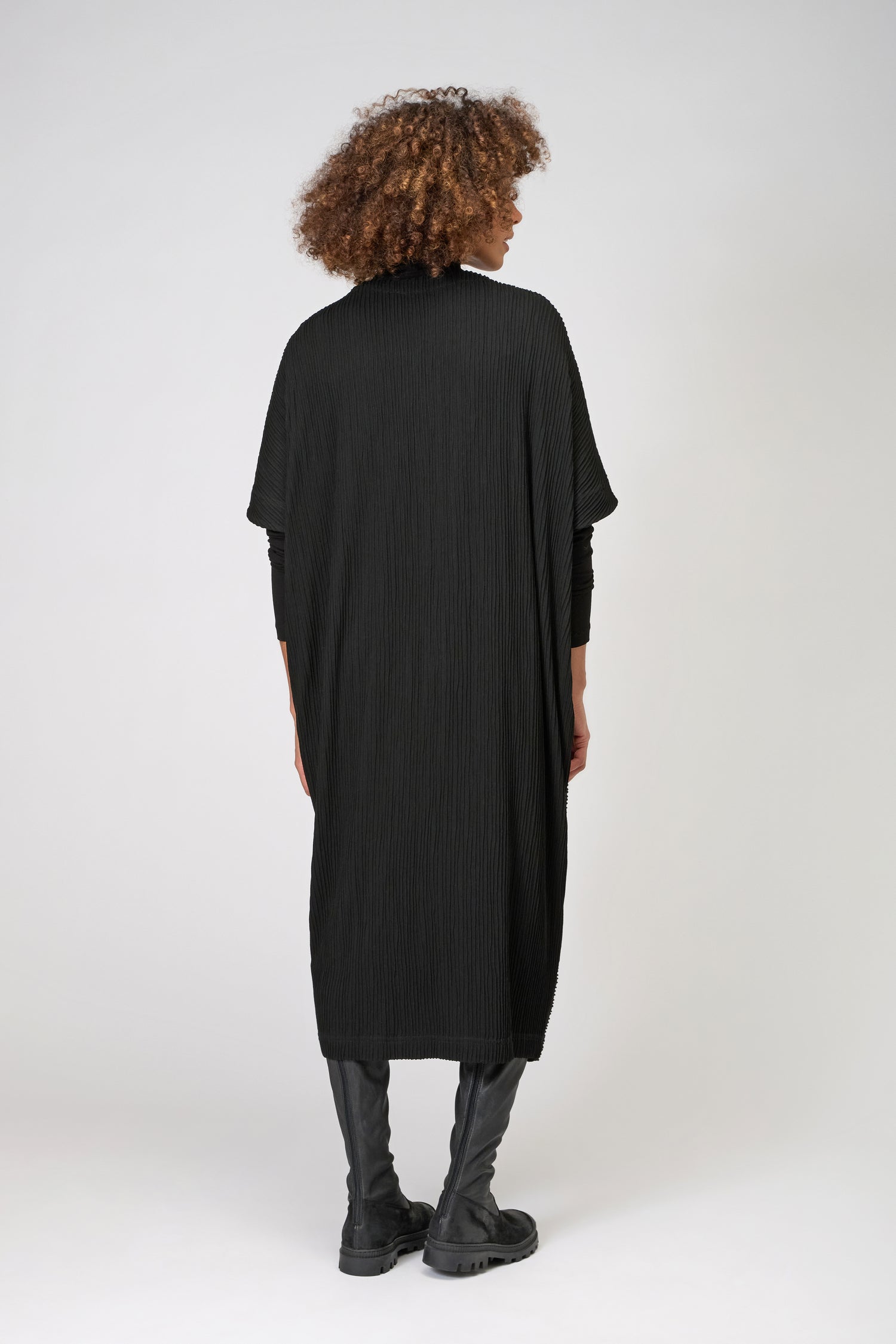 A person faces away from the camera, wearing the Textured Wave Jersey Dress—a long black, kaftan-inspired pleat-effect viscose jersey piece—paired with black pants and boots against a plain light background.