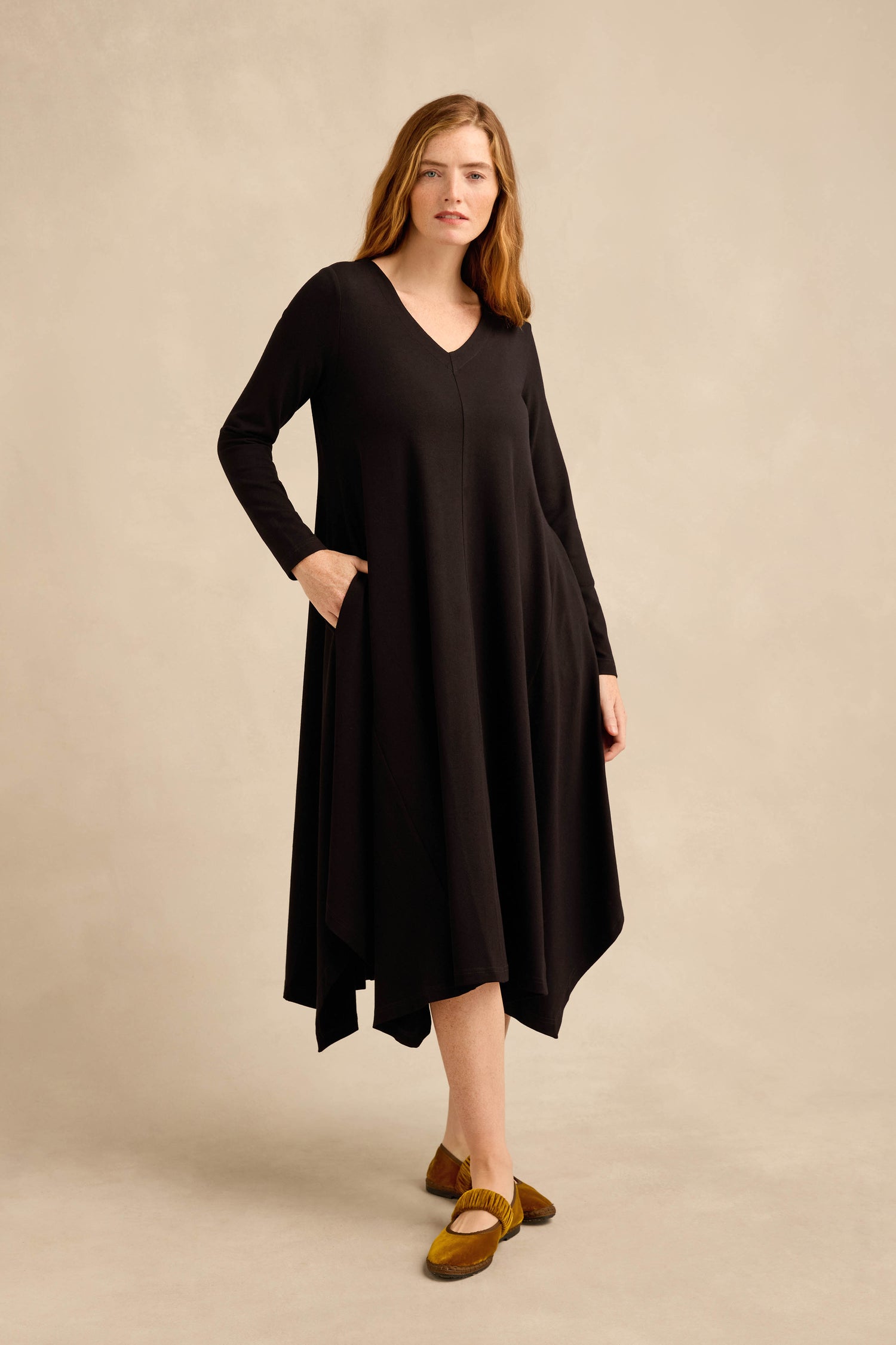 A woman stands in a studio against a neutral beige background, wearing the Fluid Crepe Flared Dress— a loose-fitting black dress with long sleeves and pockets—paired with brown shoes.