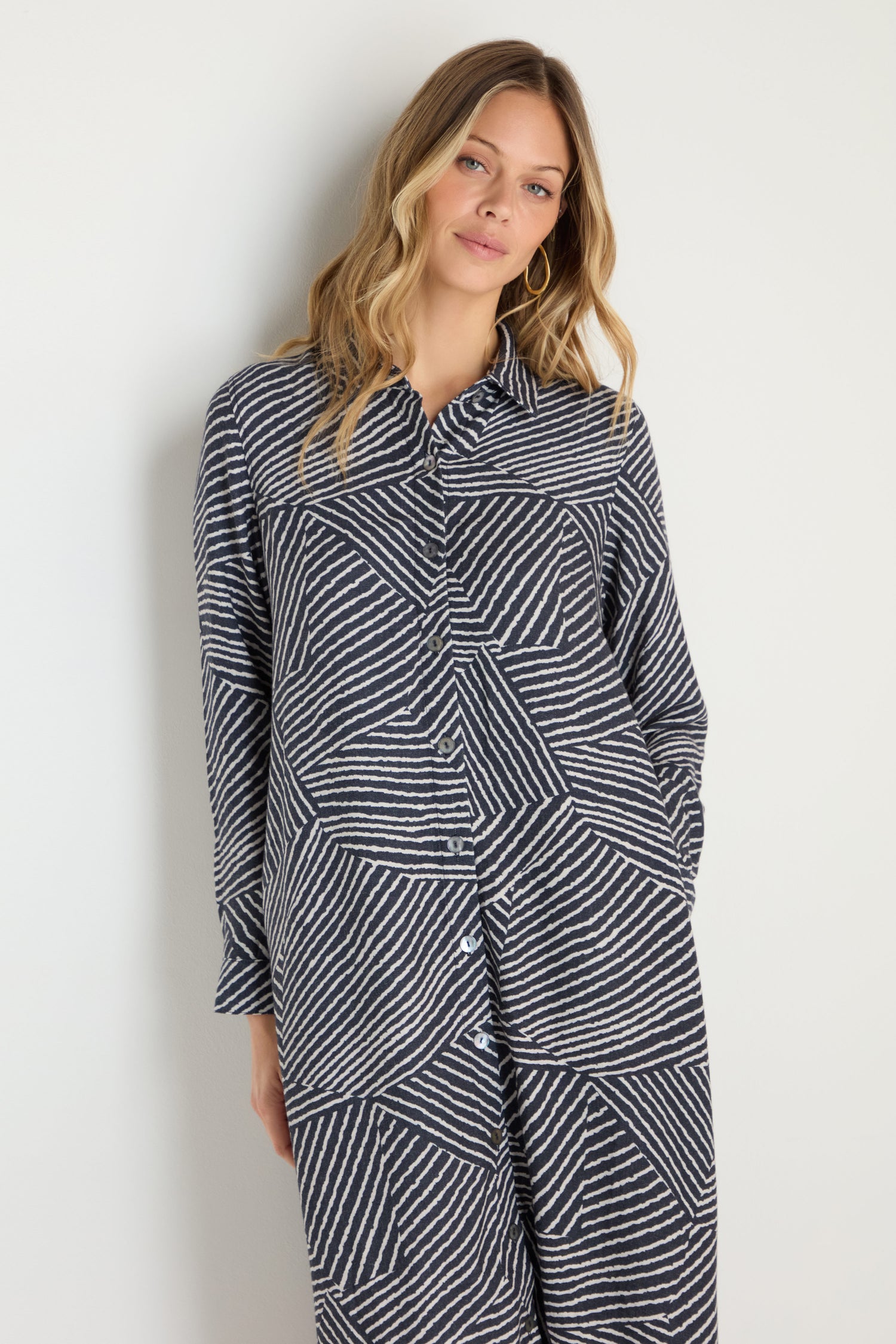 A woman stands by a plain wall wearing the Linear Prism Shirt Dress, a long-sleeve navy and white geometric print piece made in England, styled with loose hair and minimal accessories.