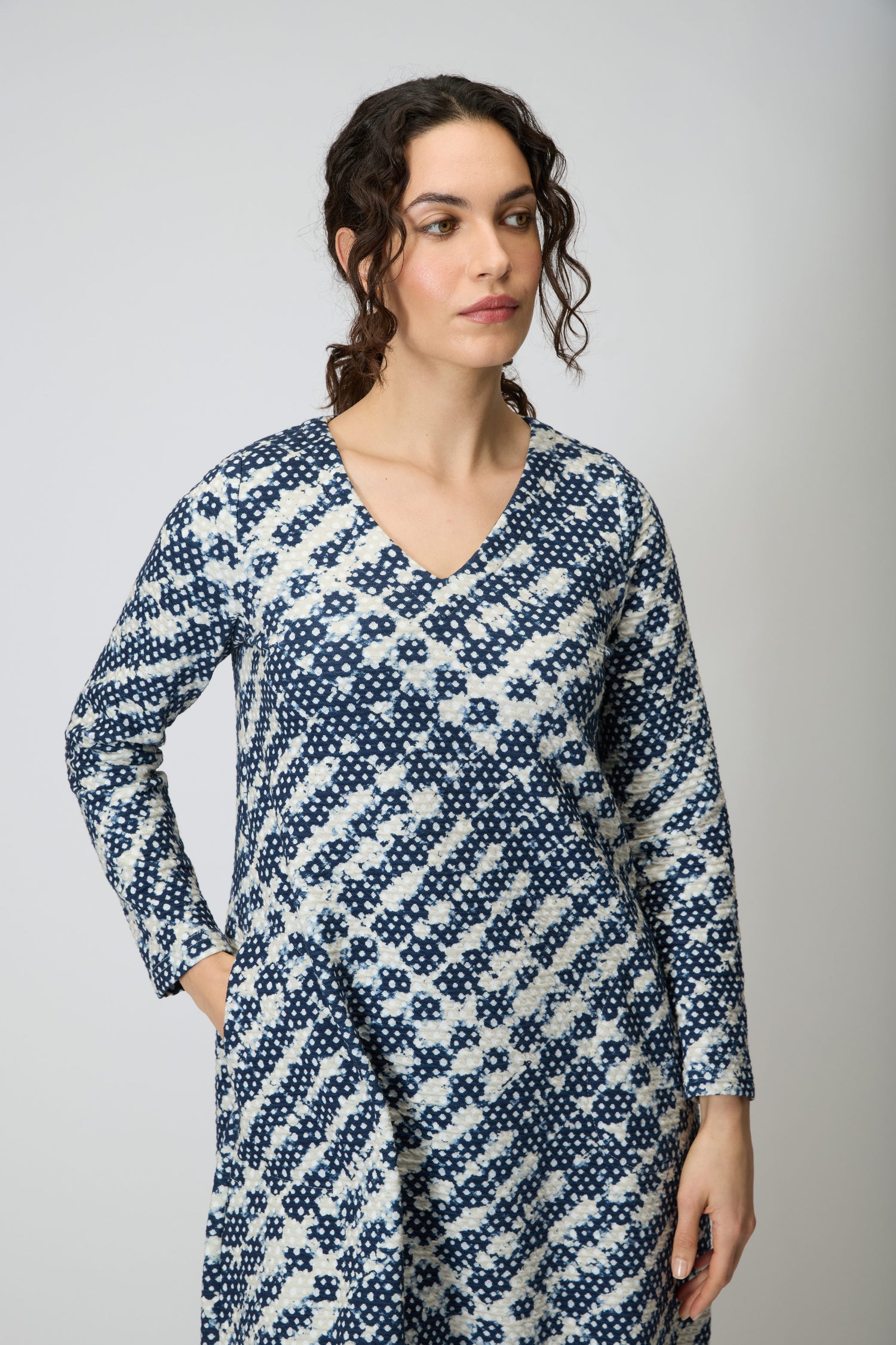 A woman with curly hair wears the Diamond Tie Dye Jersey Bubble Dress in blue and white, made of viscose jersey, featuring a bubble silhouette, standing against a plain light gray background.