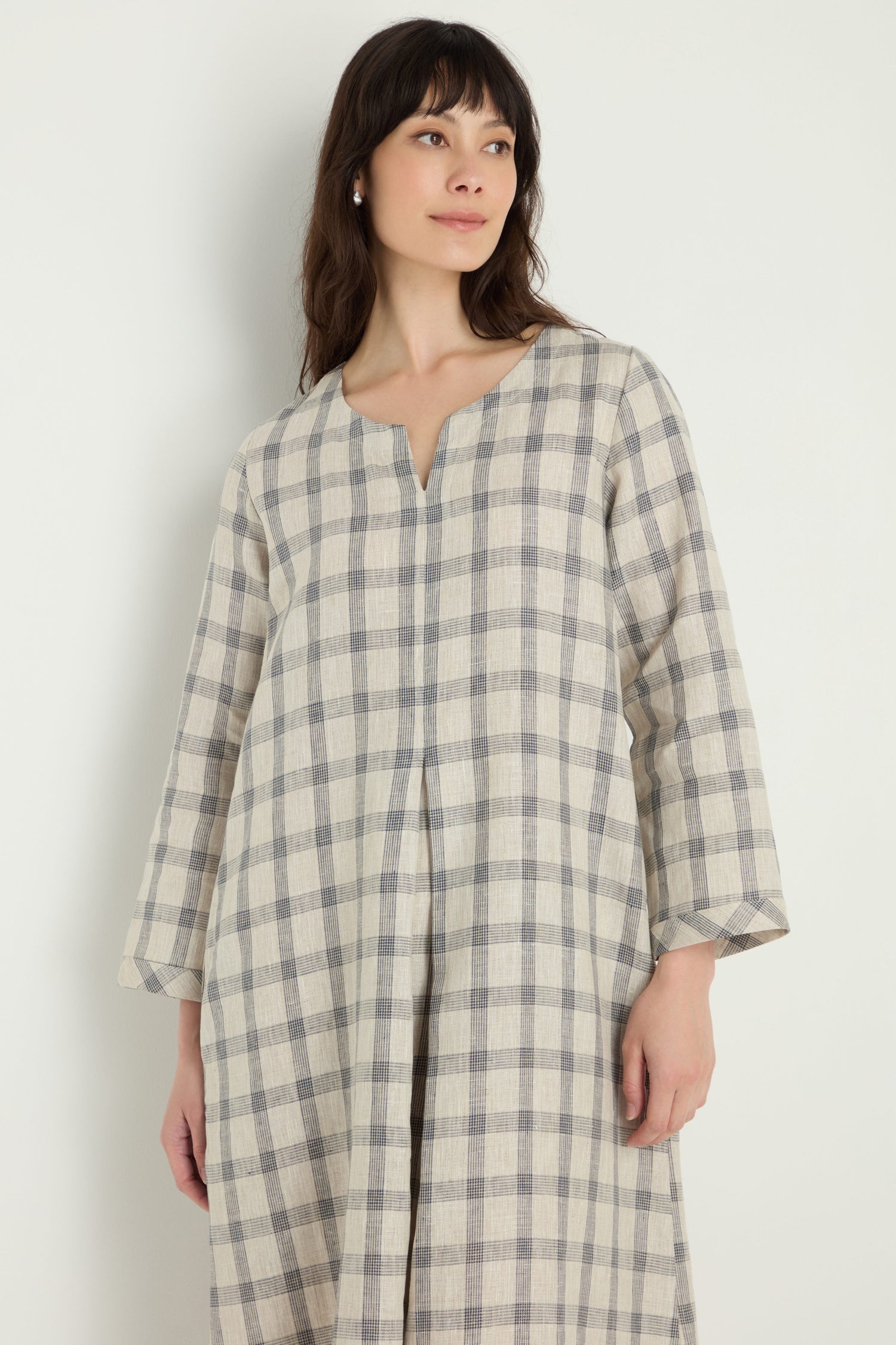 A woman stands against a plain light background wearing the Grid Check Linen Dress, featuring long sleeves, a beige and grey check pattern, and a notched neckline.