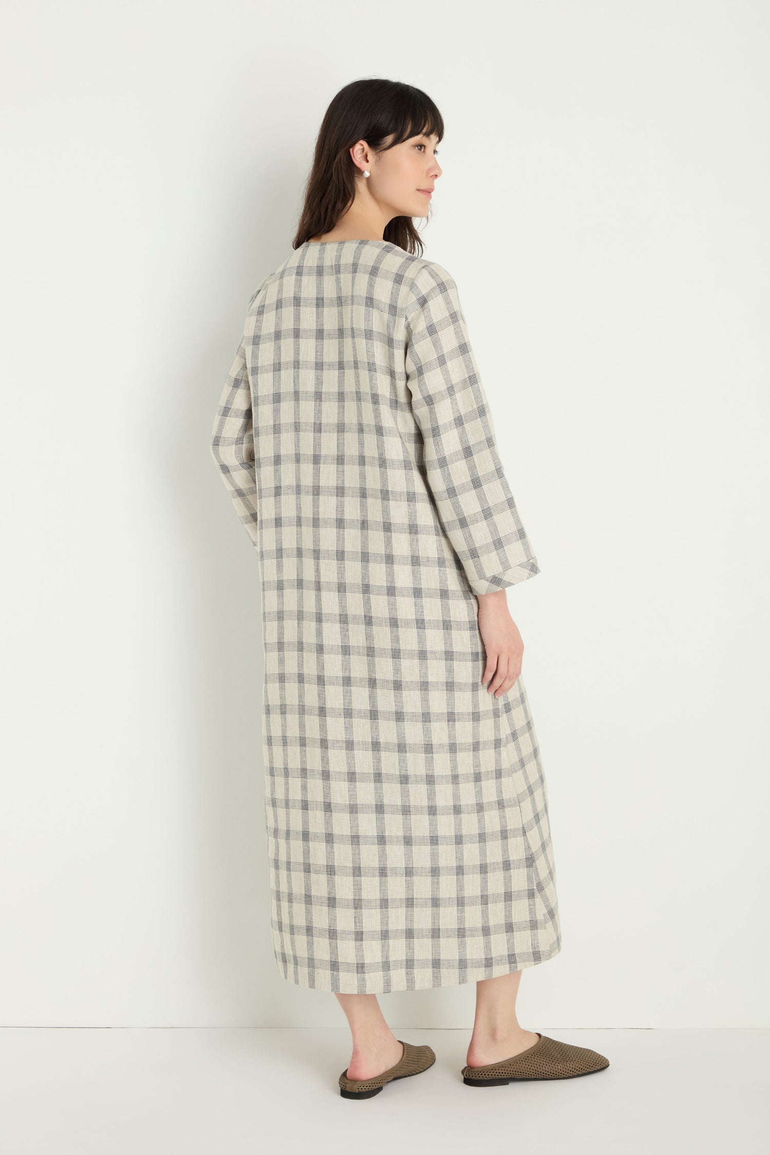 A person faces away, wearing the Grid Check Linen Dress with long sleeves and a relaxed fit in a grid pattern, paired with brown slippers, set against a plain white background.