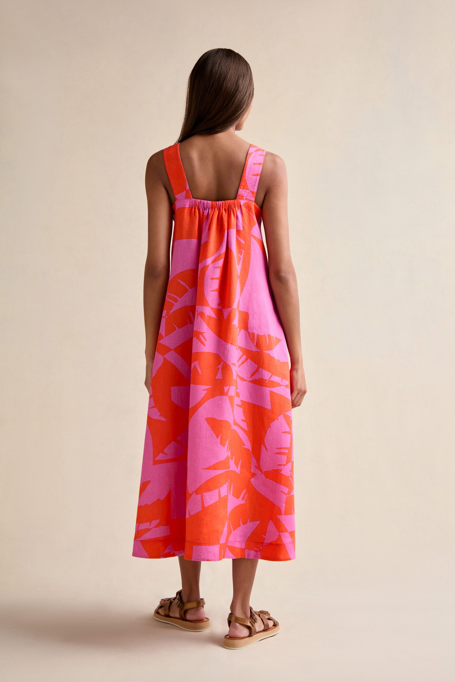 A woman faces away, wearing the Palm Mirage Linen Flared Dress—sleeveless, ankle-length with a pink and orange palm print—paired with flat brown sandals against a plain background.