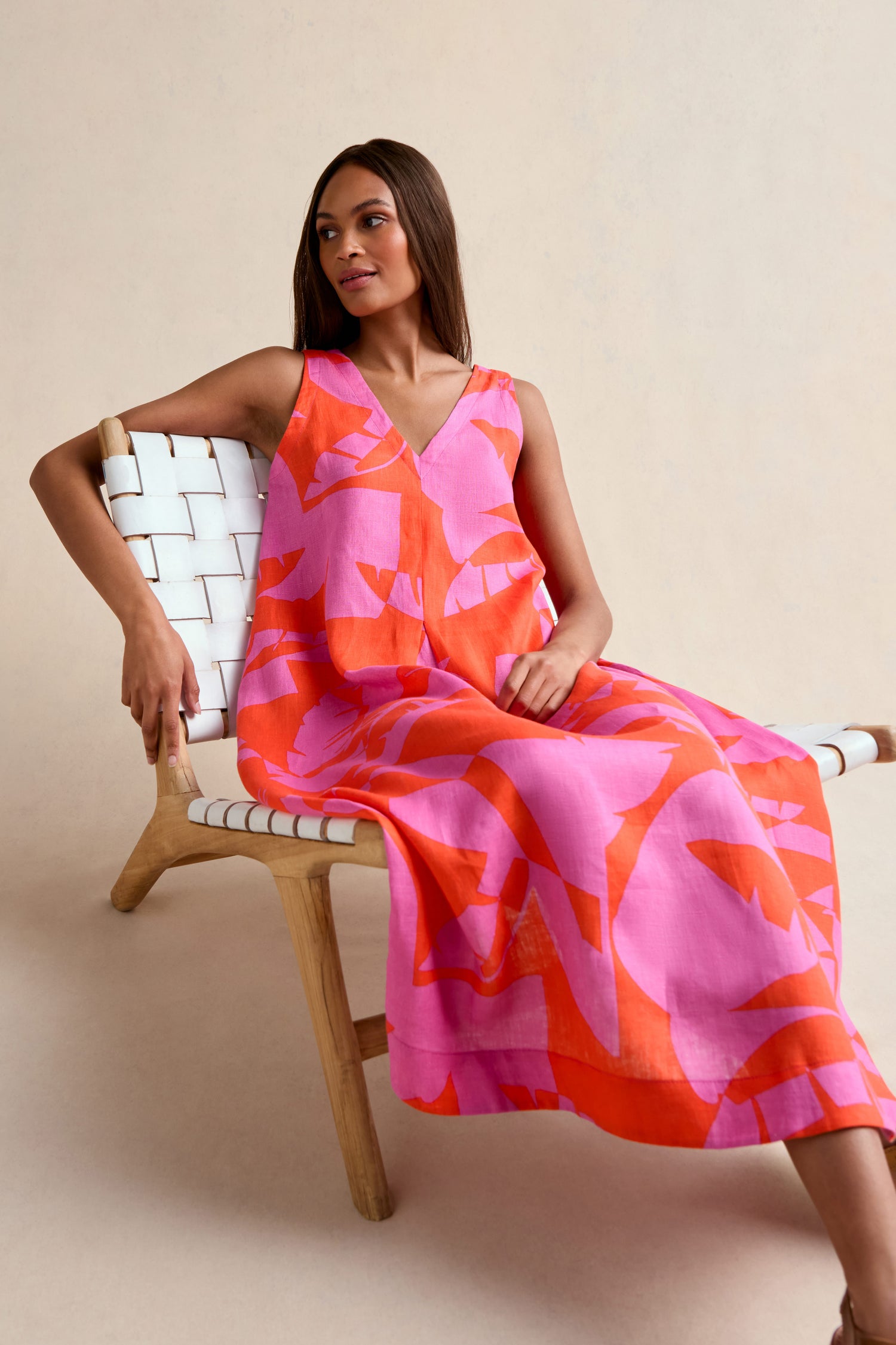 A woman sits on a wooden chair, wearing the Palm Mirage Linen Flared Dress in pink and orange patterns, gazing to the side against a light background.