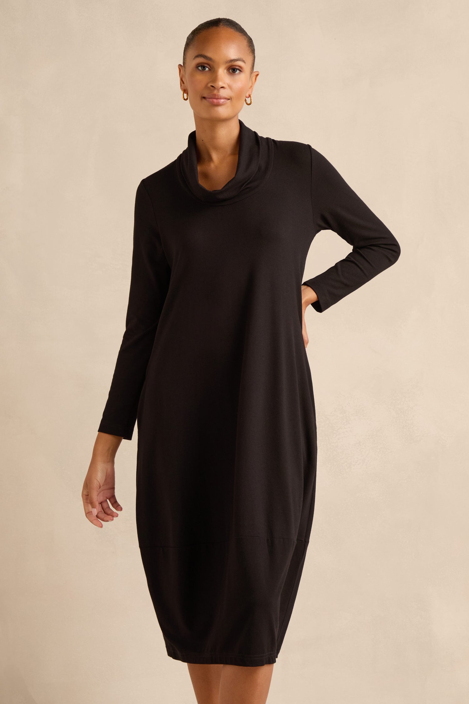 A woman wears the Fluid Crepe Bubble Dress—a long-sleeve, knee-length black bubble dress with a cowl neck—standing against a beige background.