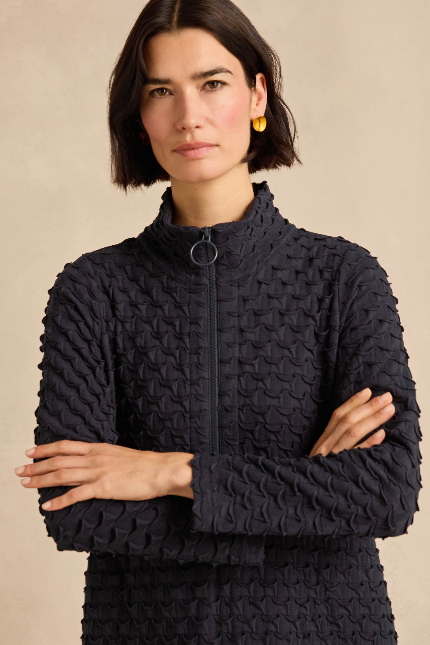A woman with short dark hair, wearing the Origami Pocket Jersey Zip Neck Bubble Dress and gold earrings, stands with her arms crossed against a neutral background.